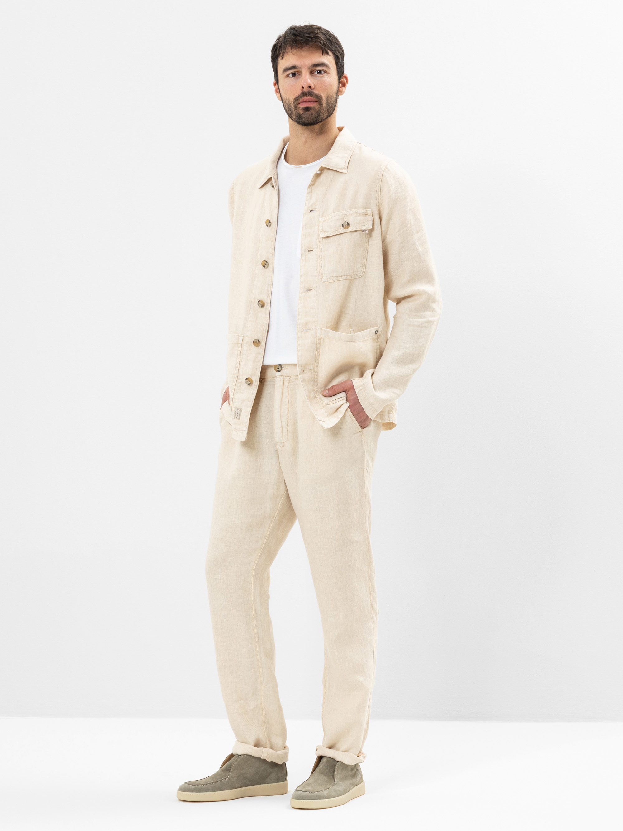 Ein Mann steht vor einem schlichten weißen Hintergrund und trägt eine hellbeige Leinenjacke und eine dazu passende Hose, ein weißes T-Shirt und hellgraue Slipper. Er hat die Hände in den Taschen und schaut selbstbewusst in die Kamera.