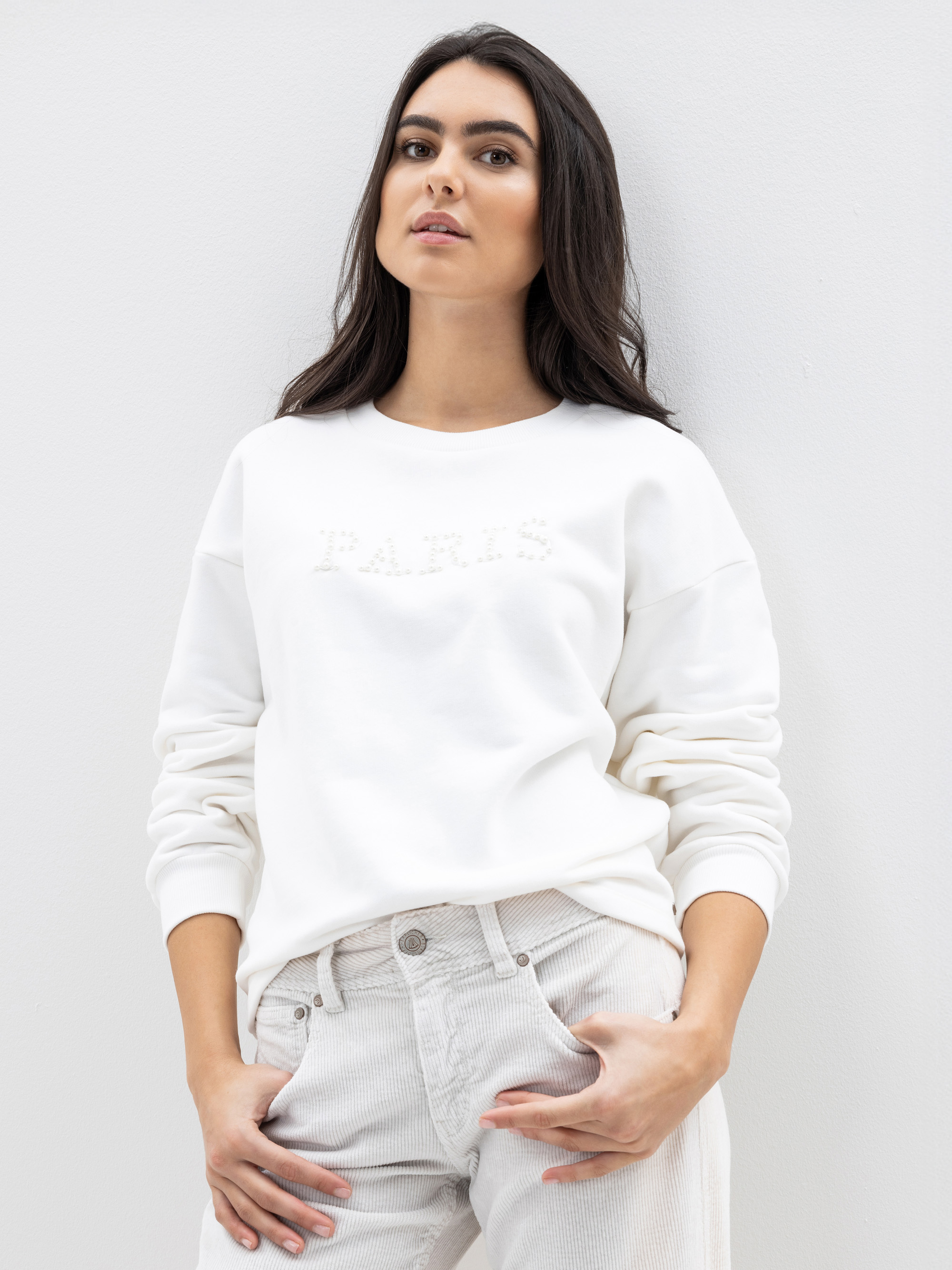 Eine Frau mit langen dunklen Haaren trägt ein weißes Sweatshirt mit der Aufschrift PARIS und helle Jeans. Sie steht vor einer schlichten weißen Wand und schaut selbstbewusst in die Kamera, die Hände in den Taschen.