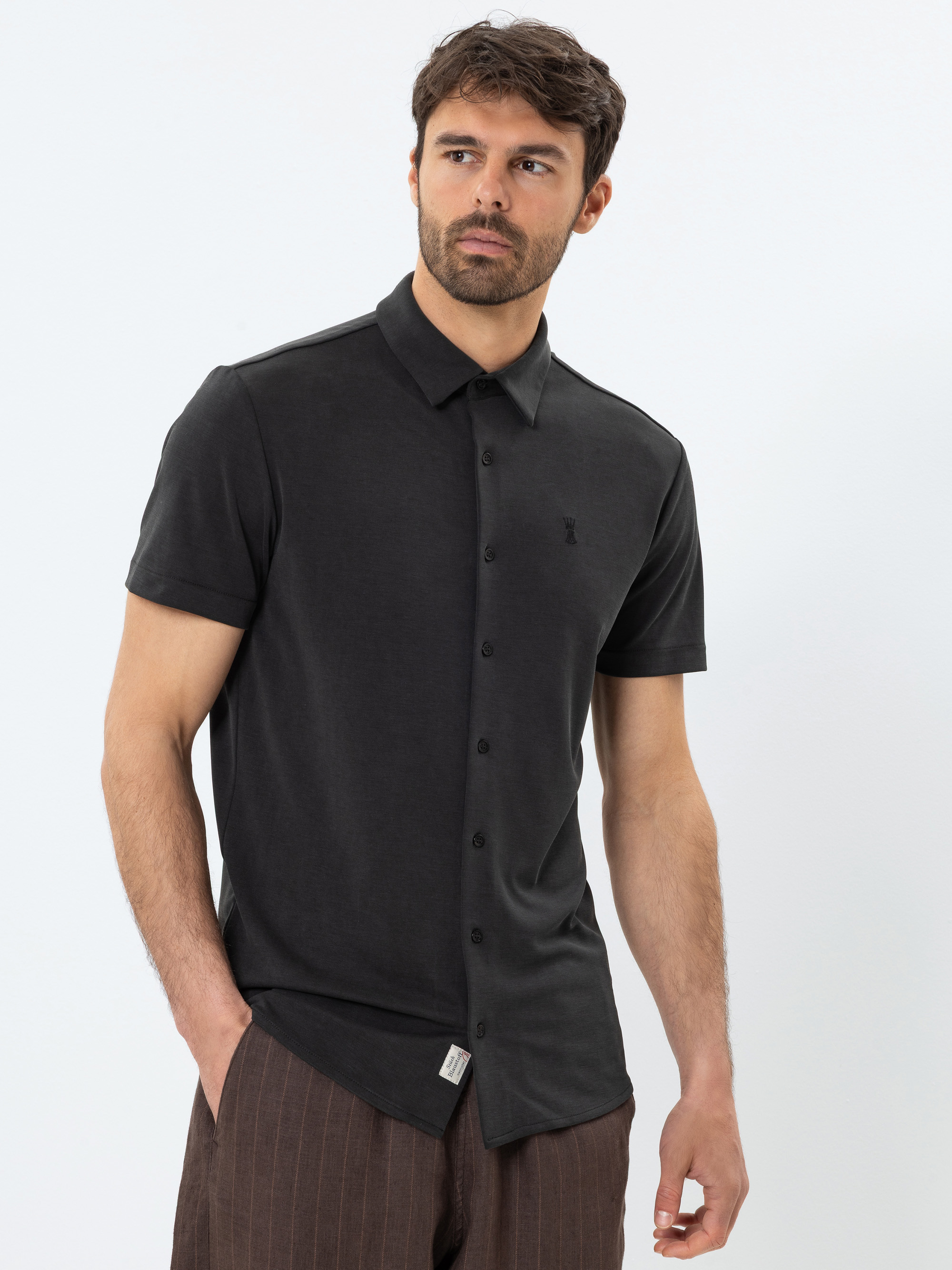 Ein Mann mit kurzen dunklen Haaren und Bart trägt ein schwarzes, kurzärmeliges Button-up-Hemd und eine braune Nadelstreifenhose. Er steht vor einem schlichten weißen Hintergrund und hat eine Hand in der Tasche.