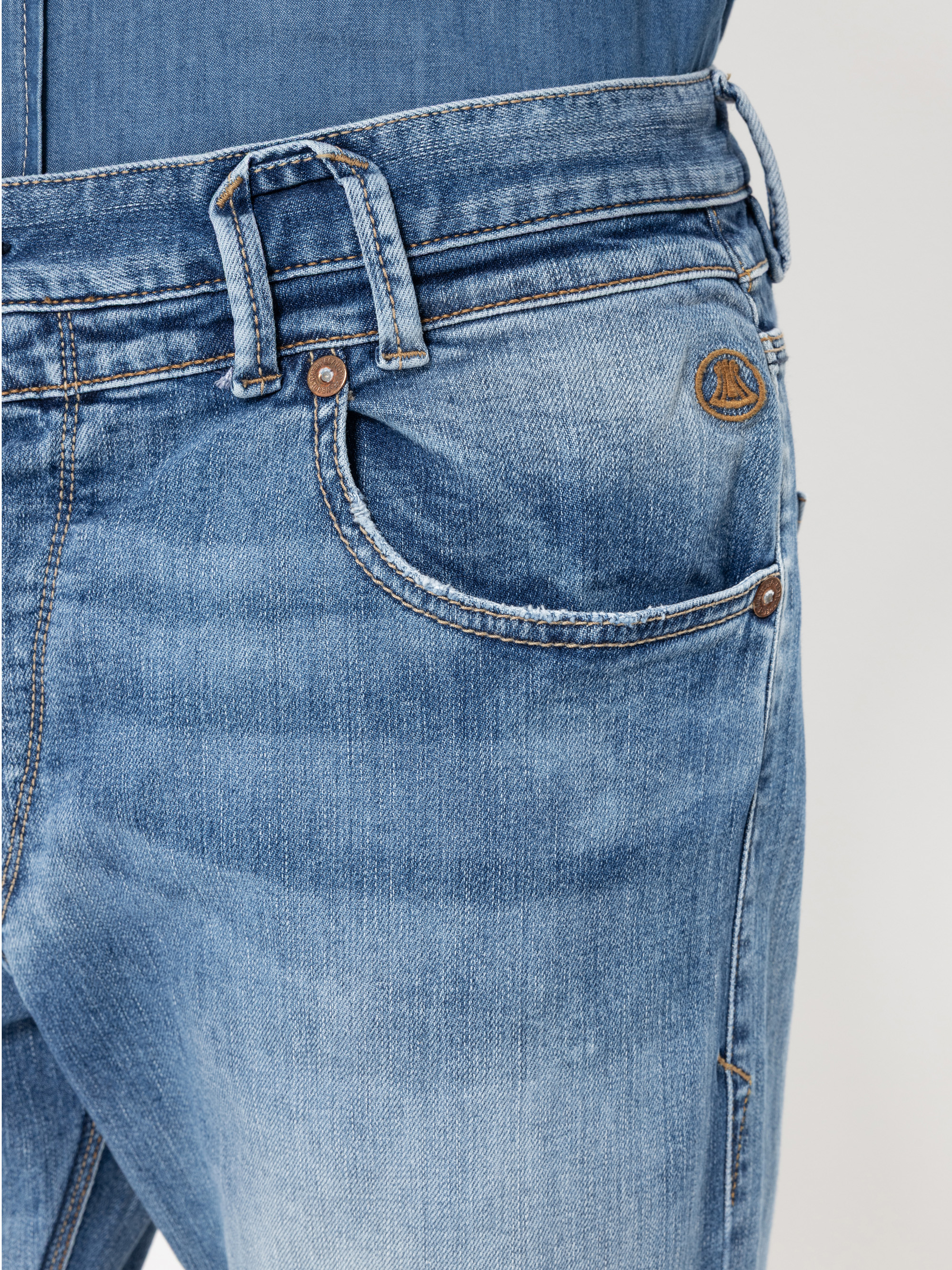 Nahaufnahme einer hellblauen Denim-Jeans mit Vordertasche, Gürtelschlaufen und einem kleinen runden braunen Logoaufnäher über der Tasche. Der Stoff weist verblichene und leicht beanspruchte Details auf.