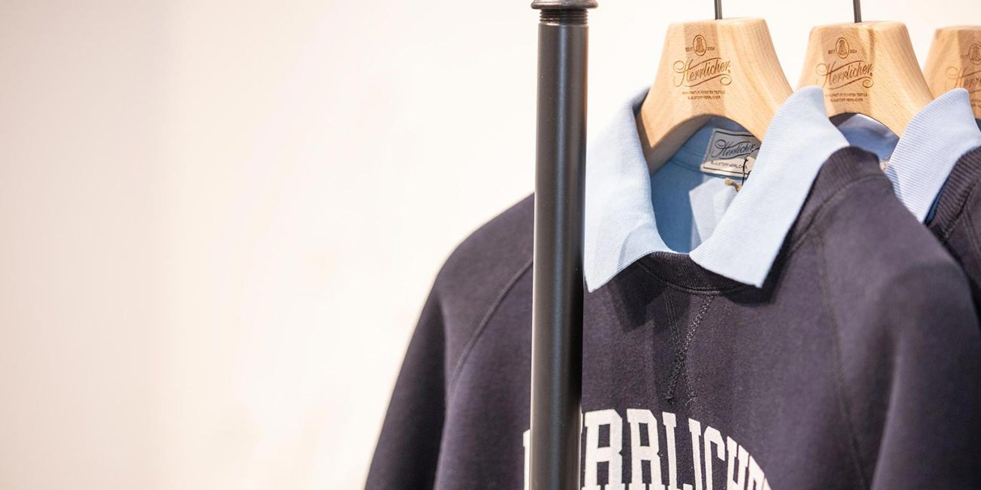 Nahaufnahme von marineblauen Sweatshirts mit hellblauen Kragen, die auf Holzbügeln an einem Kleiderständer vor einem hellen Hintergrund hängen. Die Sweatshirts haben einen weißen Text auf der Brust.