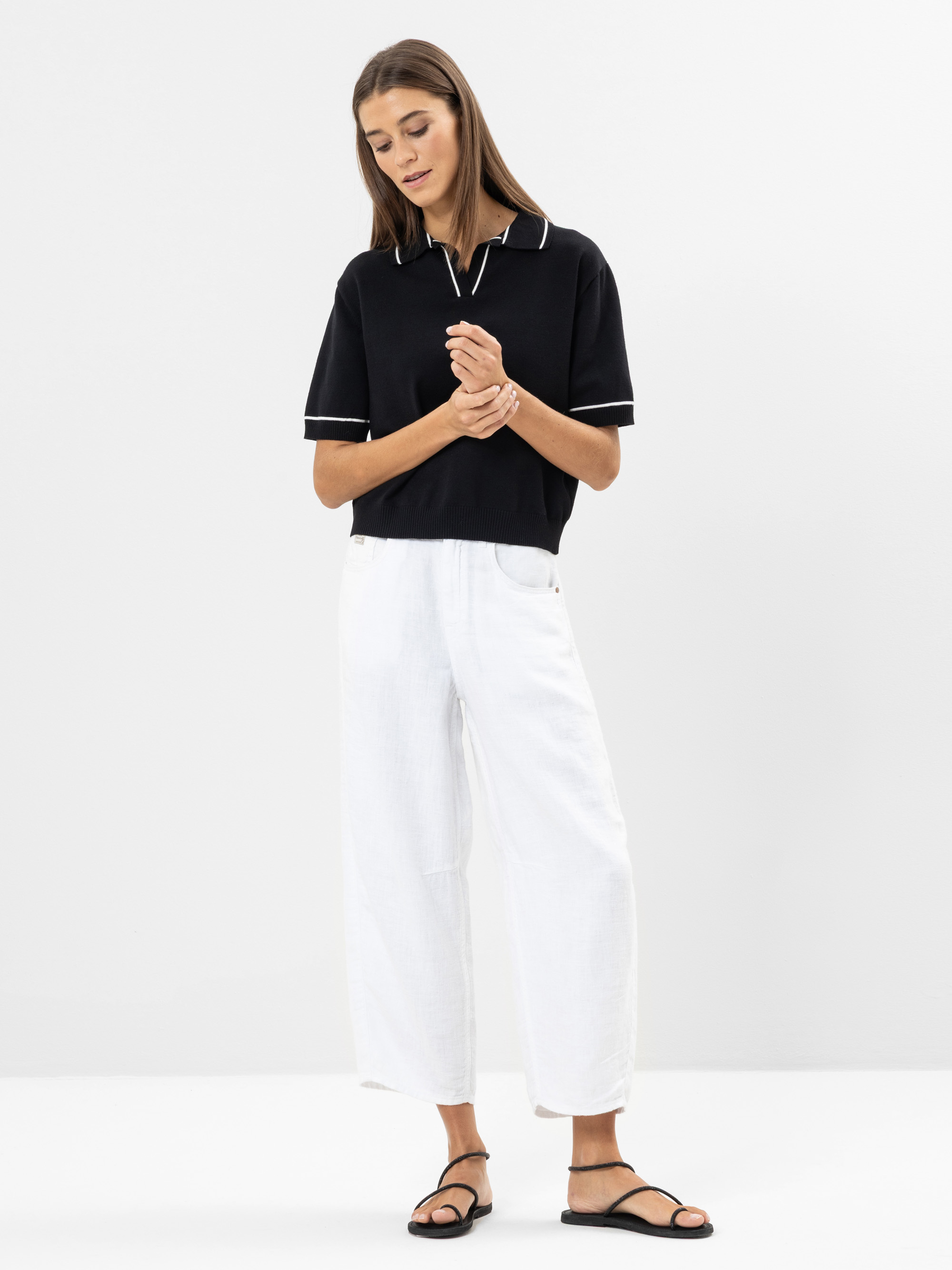 Eine Frau steht vor einem schlichten weißen Hintergrund. Sie trägt ein schwarzes kurzärmeliges Poloshirt mit weißem Rand, eine weite weiße Hose und schwarze Sandalen. Sie blickt nach unten, die Hände sanft vor sich verschränkt.