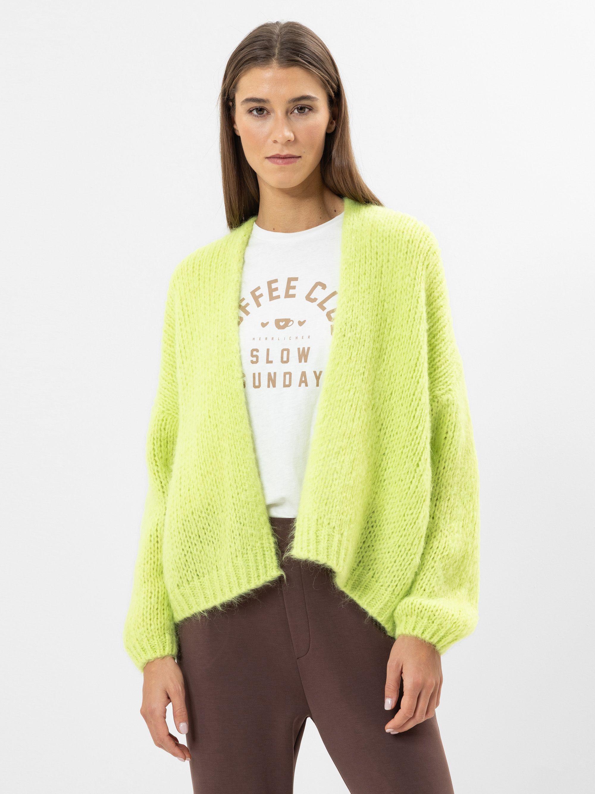 Eine Frau mit glattem braunem Haar trägt eine lockere, leuchtend gelb-grüne Strickjacke über einem weißen grafischen T-Shirt und einer braunen Hose und steht vor einem schlichten weißen Hintergrund.