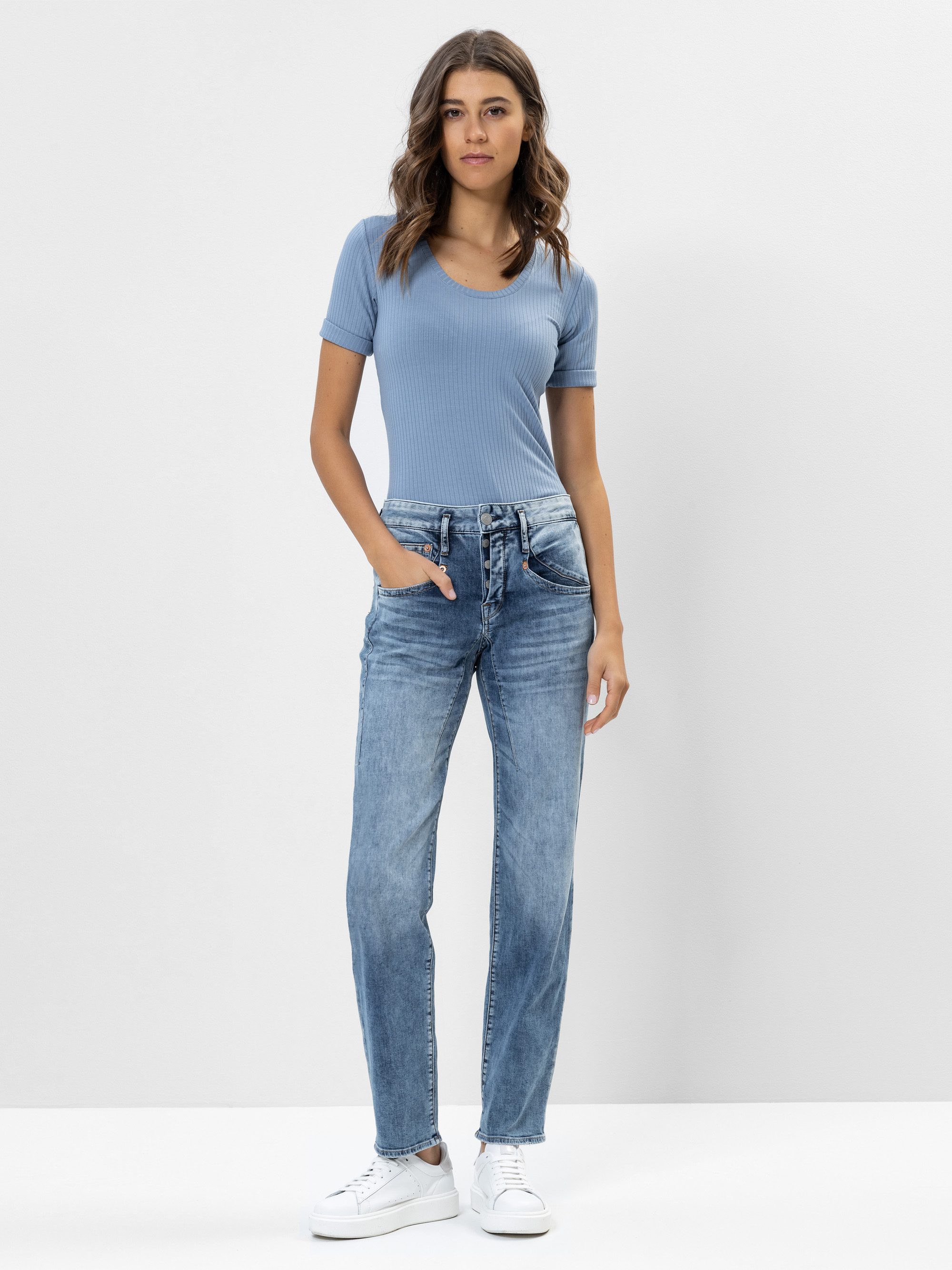 Eine Frau steht vor einem schlichten weißen Hintergrund und trägt ein hellblaues, kurzärmeliges, geripptes Oberteil mit V-Ausschnitt, blaue Jeans mit geradem Bein und weiße Turnschuhe. Sie hat gewelltes braunes Haar und einen entspannten Gesichtsausdr