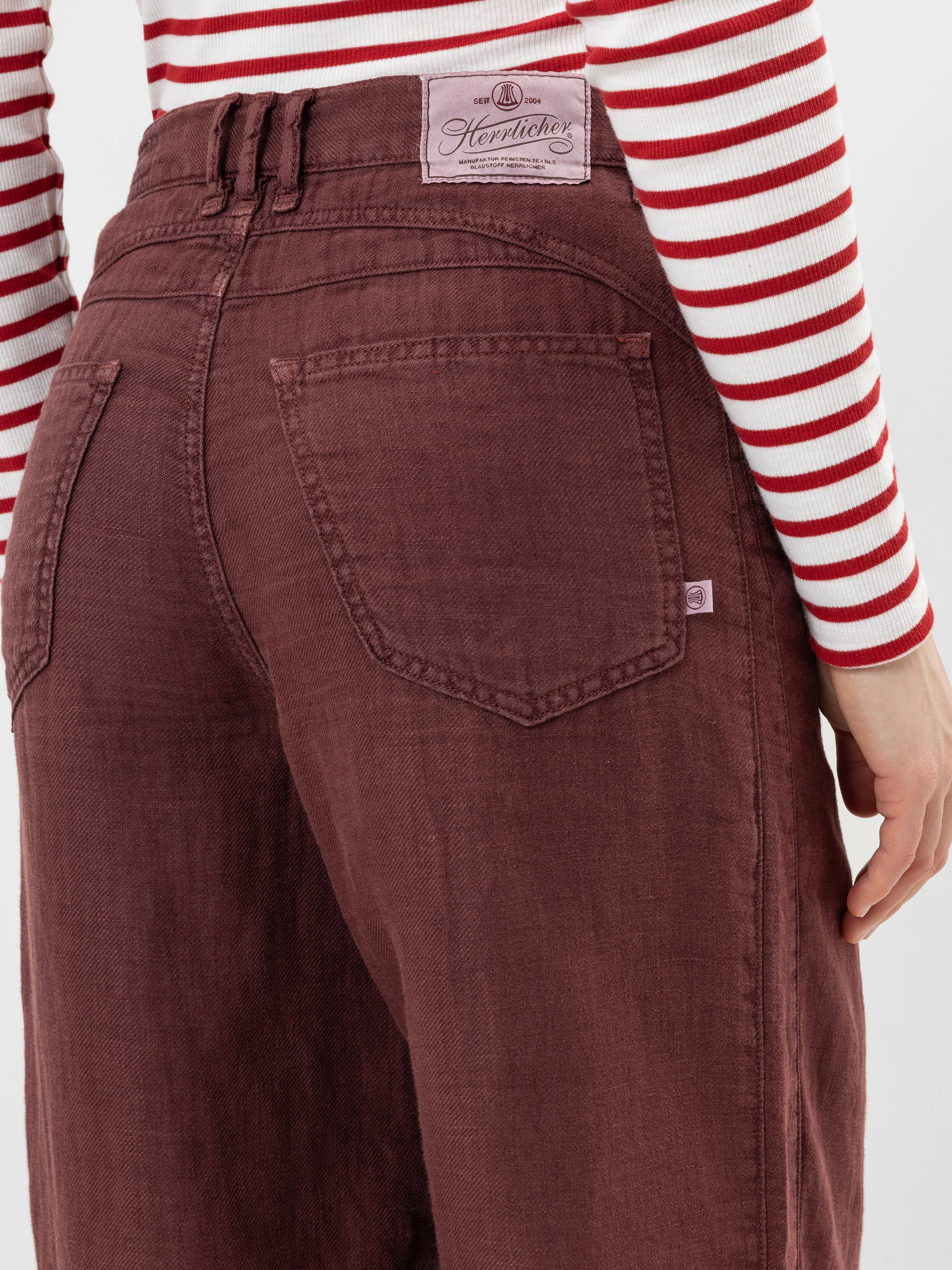 Eine Person trägt eine burgunderfarbene Jeans mit hoher Taille, sichtbaren Gesäßtaschen und einem Etikett, dazu ein rot-weiß gestreiftes langärmeliges Hemd. Das Bild konzentriert sich auf die Rückenansicht der Jeans.