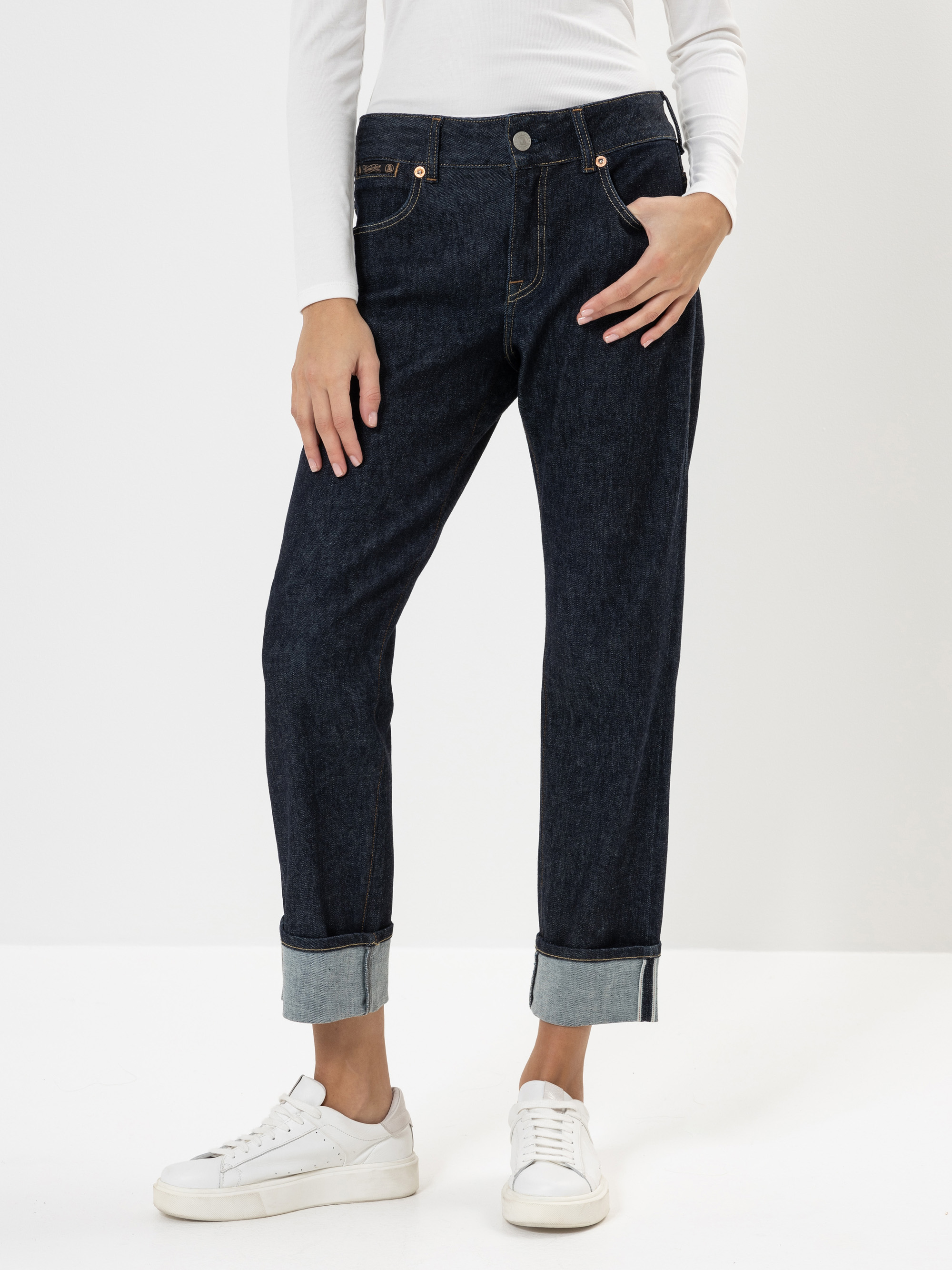 Eine Person, die dunkelblaue Jeans mit Bündchen, ein weißes langärmeliges Oberteil und weiße Turnschuhe trägt, steht vor einem schlichten weißen Hintergrund und hat eine Hand in der Tasche.