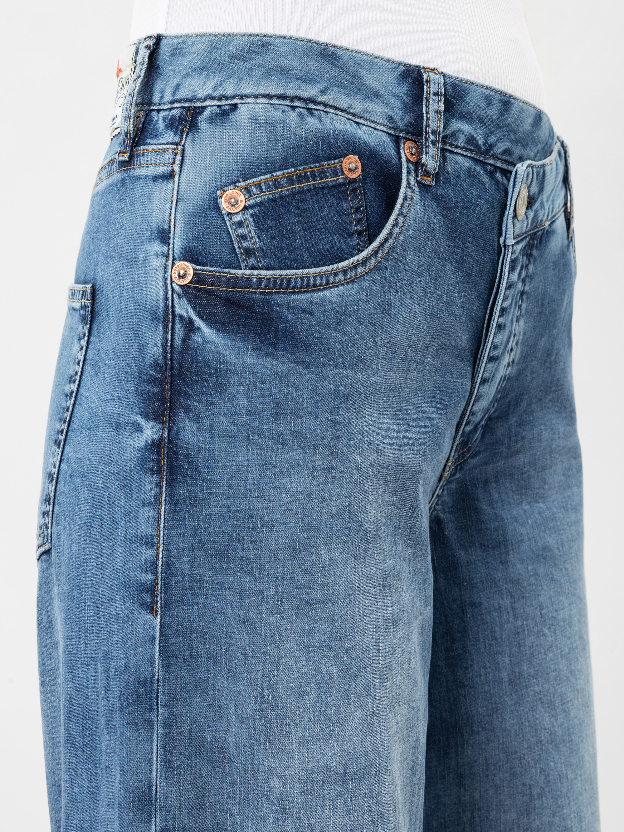 Nahaufnahme einer Person, die eine blaue Denim-Jeans mit Kupfernieten und Vordertaschen trägt, gepaart mit einem weißen Hemd zum Hineinstecken. Der Fokus liegt auf der Seite und der Vorderseite der Jeans vor einem schlichten Hintergrund.