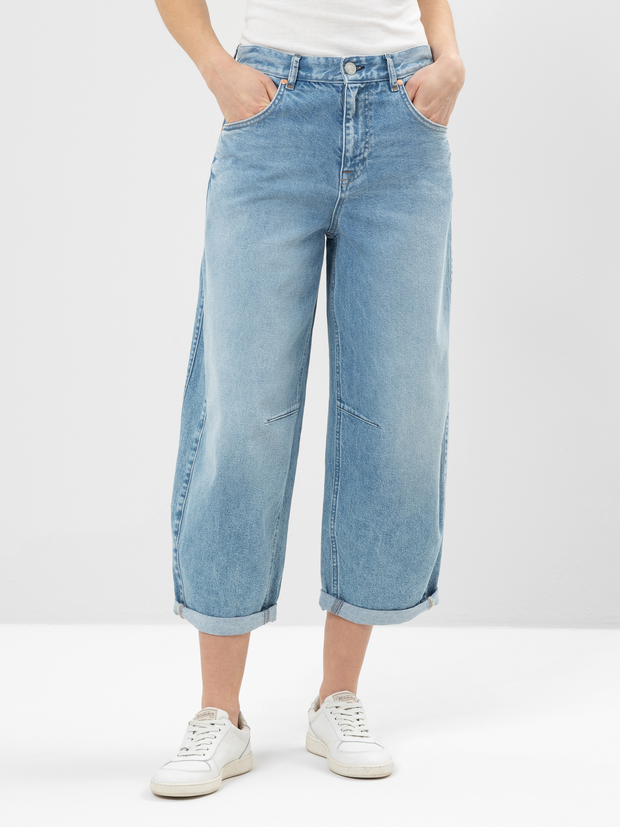 Eine Person, die hellblaue, weit geschnittene Jeans mit aufgerollten Bündchen, ein weißes Oberteil und weiße Turnschuhe trägt, steht vor einem schlichten hellen Hintergrund, die Hände in den Taschen.