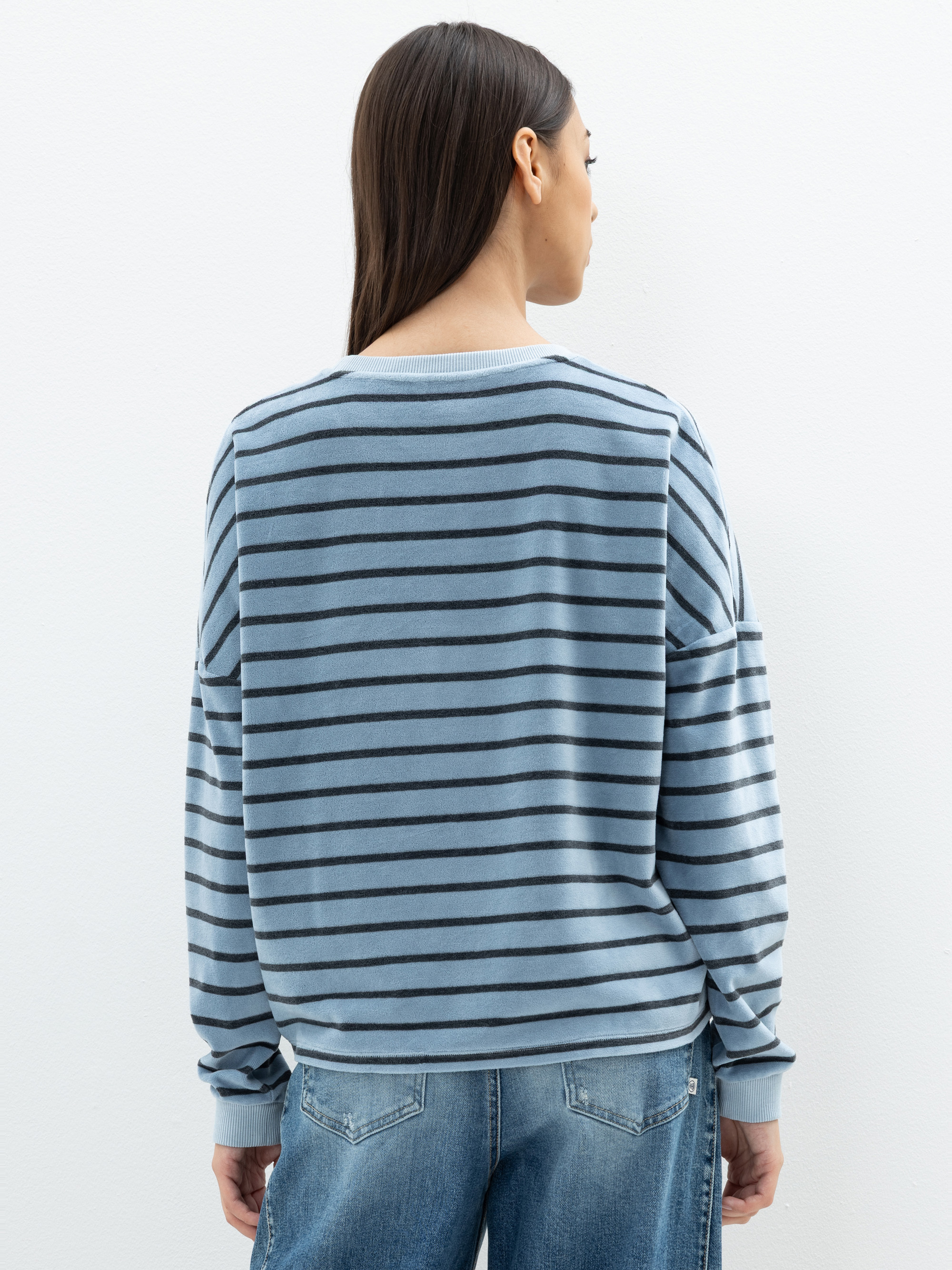 Eine Frau mit langen braunen Haaren, von hinten gesehen, trägt einen locker sitzenden blau-schwarz gestreiften Pullover und blaue Jeans und steht vor einer schlichten weißen Wand.