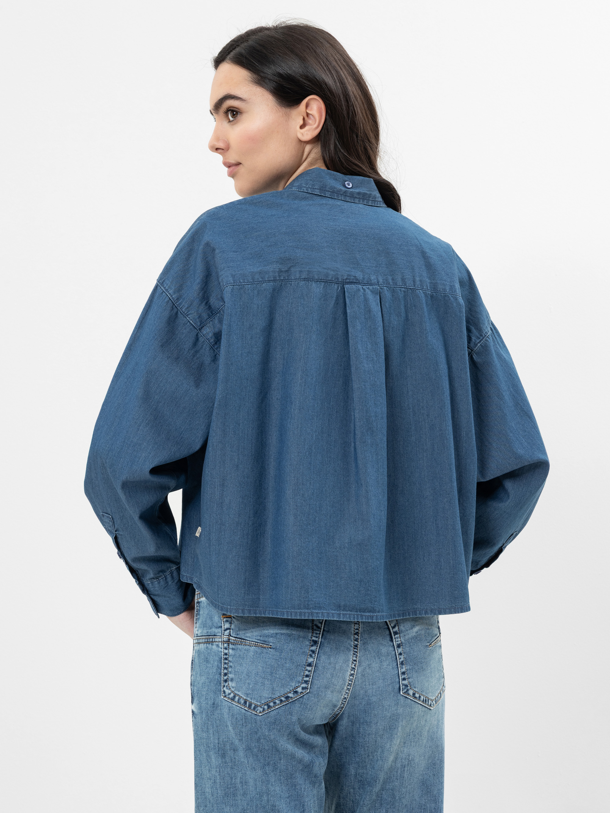 Eine Frau mit langen dunklen Haaren, die ein locker sitzendes blaues Jeanshemd und blaue Jeans trägt, steht mit dem Rücken zur Kamera vor einem schlichten weißen Hintergrund. Ihre Hände stecken in den Gesäßtaschen.
