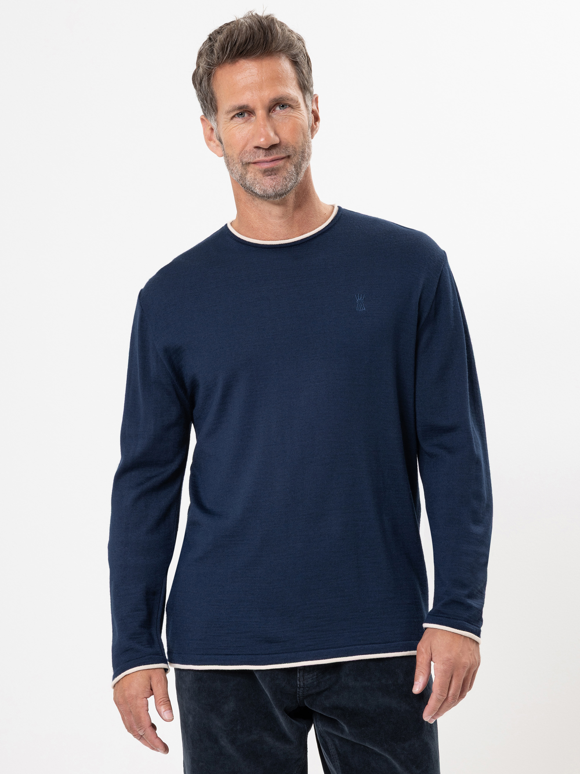 Ein Mann mit kurzem, grauem Haar und Bart trägt ein marineblaues, langärmeliges Hemd mit weißem Saum, steht vor einem schlichten weißen Hintergrund und lächelt leicht.