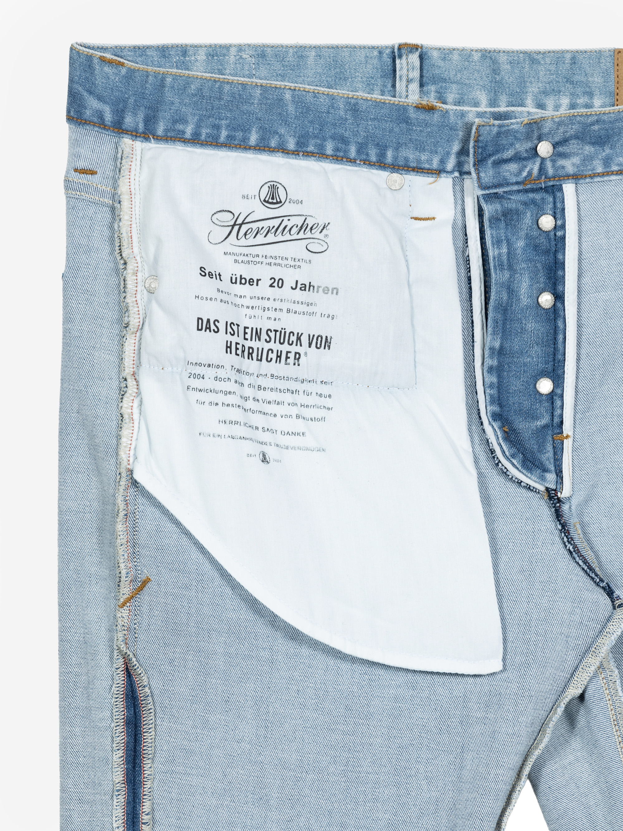 Nahaufnahme der Innenseite einer hellblauen Jeans mit aufgedrucktem Branding und Text auf dem Taschenfutter, Metallknöpfen und sichtbaren Nähten.