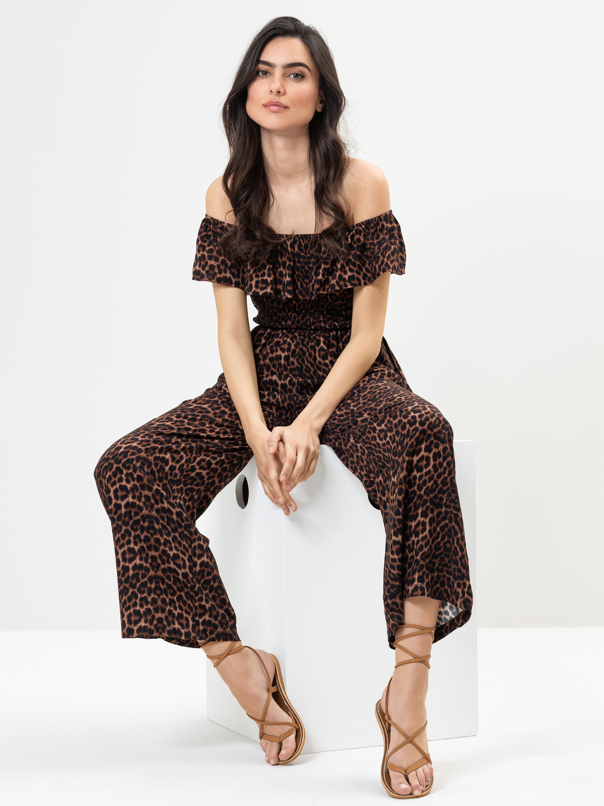 Frau mit langen dunklen Haaren, die einen schulterfreien braun-schwarzen Jumpsuit mit Leopardenmuster und Riemchensandalen trägt, sitzt auf einem weißen Block vor einem schlichten Hintergrund. Sie blickt mit entspanntem Blick nach vorne.