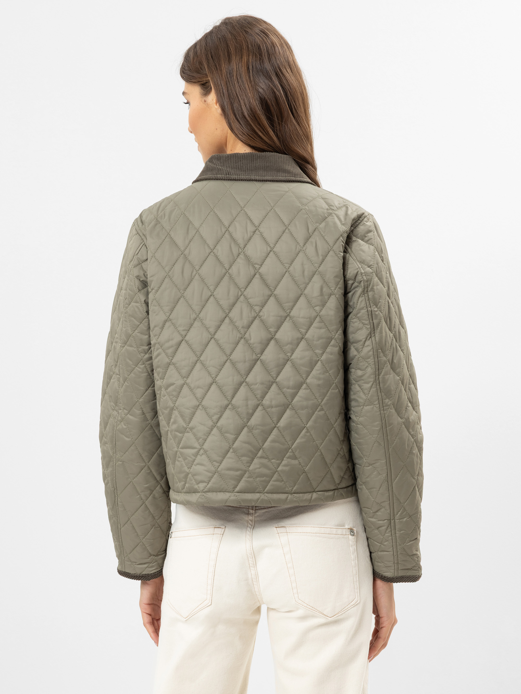 Eine Frau mit langen braunen Haaren wird von hinten gezeigt. Sie trägt eine gesteppte olivgrüne Jacke und cremefarbene Jeans vor einem schlichten hellen Hintergrund.