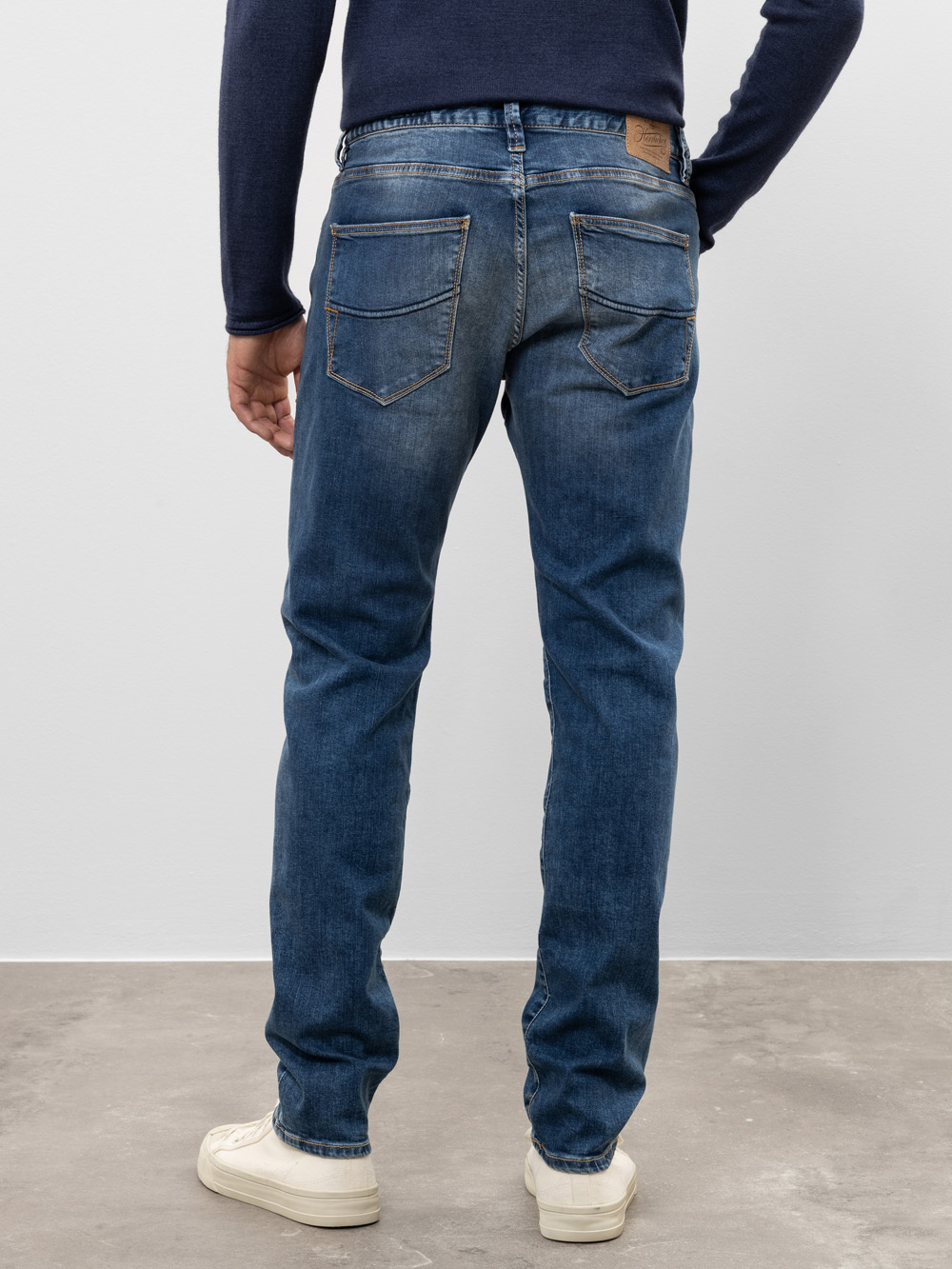 Eine Person steht mit dem Gesicht zur Wand und trägt eine blaue Jeans, ein marineblaues langärmeliges Hemd und weiße Turnschuhe. Die Umgebung ist ein Innenraum mit einer schlichten hellgrauen Wand und einem Betonboden.