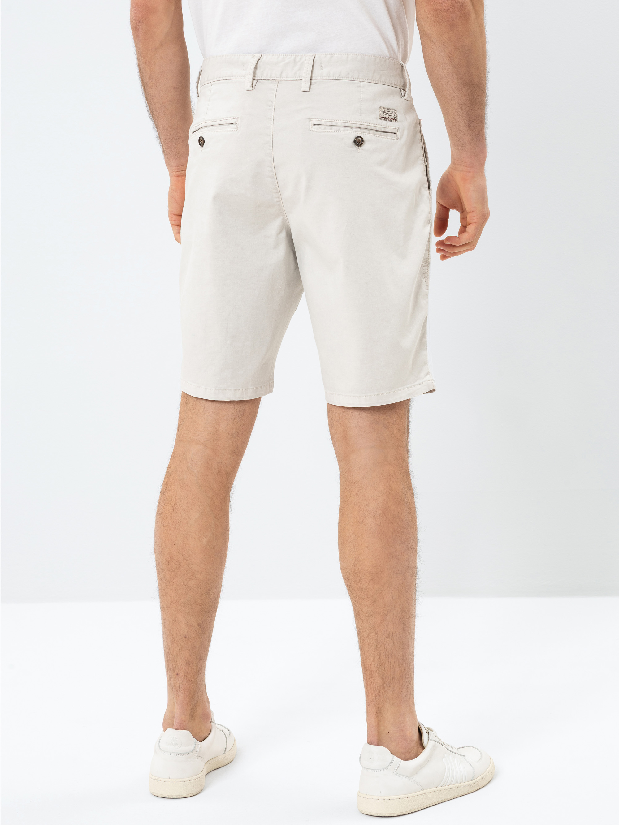 Ein Mann steht mit dem Gesicht nach hinten, trägt hellbeige Shorts, ein weißes T-Shirt und weiße Turnschuhe vor einem schlichten weißen Hintergrund. Die Shorts haben Gesäßtaschen mit Knöpfen.