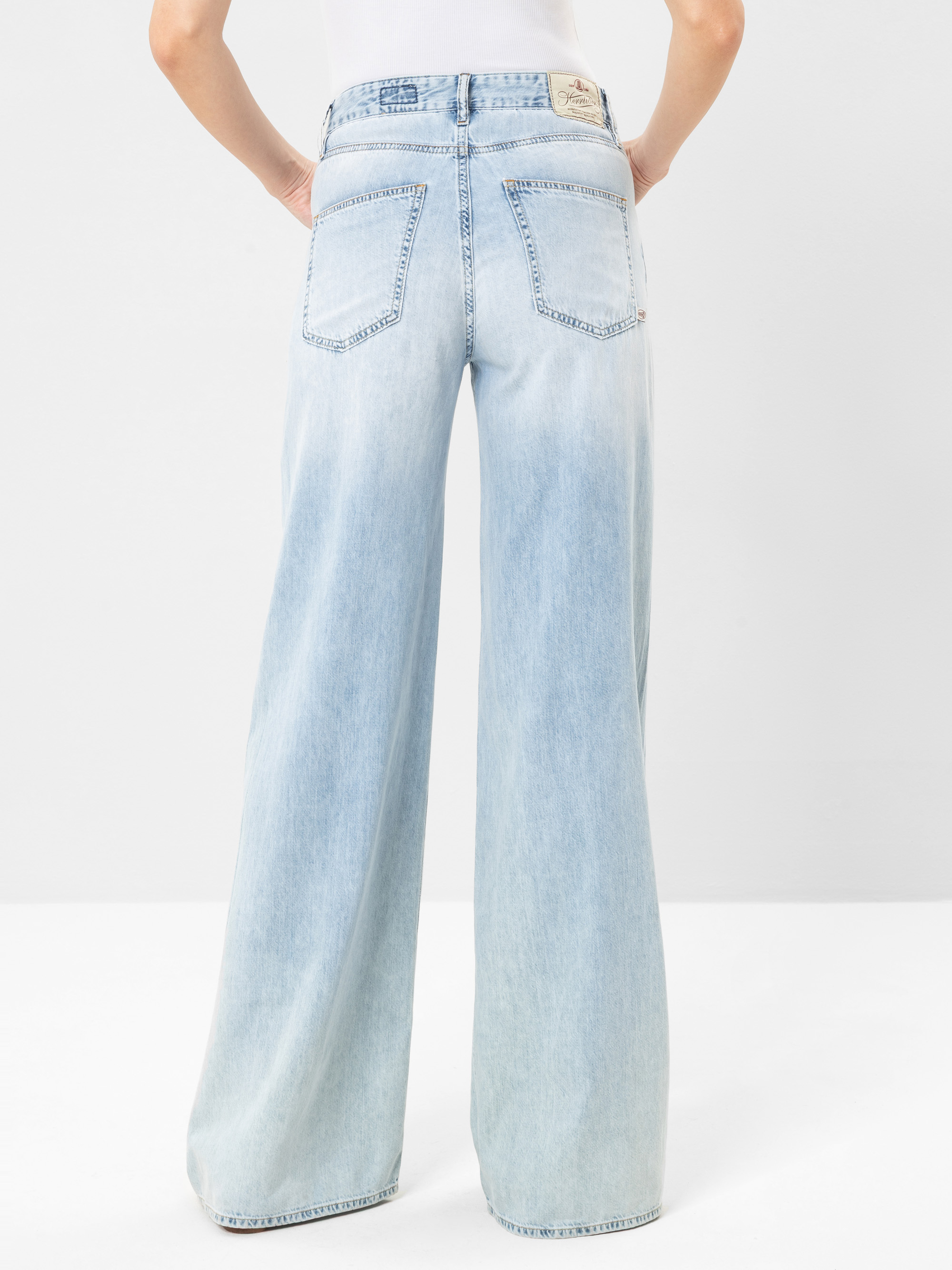 Person, die eine hoch taillierte, hell gewaschene Jeans mit weitem Bein trägt, von hinten gesehen, mit zwei großen Gesäßtaschen und einem beigen Markenaufnäher am Bund. Der Hintergrund ist einfarbig und hell gefärbt.