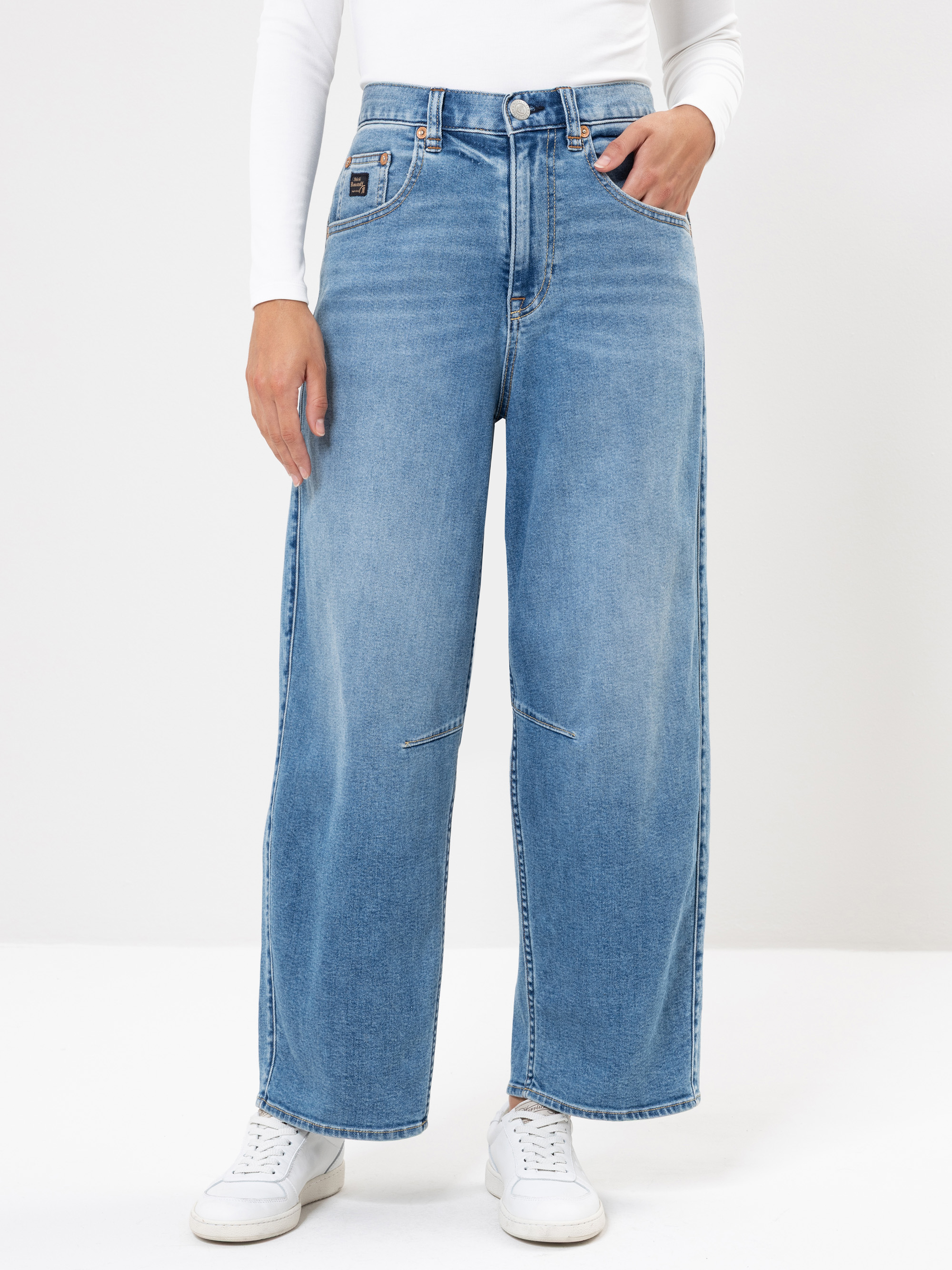 Eine Person, die hellblaue, weit geschnittene Jeans, ein weißes, langärmeliges Oberteil und weiße Turnschuhe trägt, steht mit einer Hand in der Tasche vor einem schlichten weißen Hintergrund.