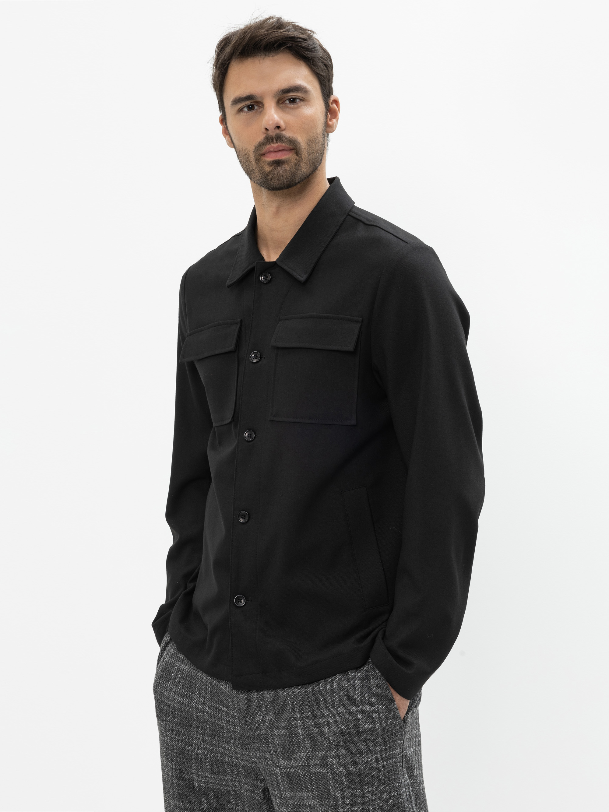 Ein Mann steht vor einem schlichten weißen Hintergrund. Er trägt ein schwarzes Button-up-Hemd mit zwei Brusttaschen und eine grau karierte Hose. Er trägt einen gestutzten Bart und Schnurrbart, hat die Hände in den Taschen und schaut selbstbewusst in d
