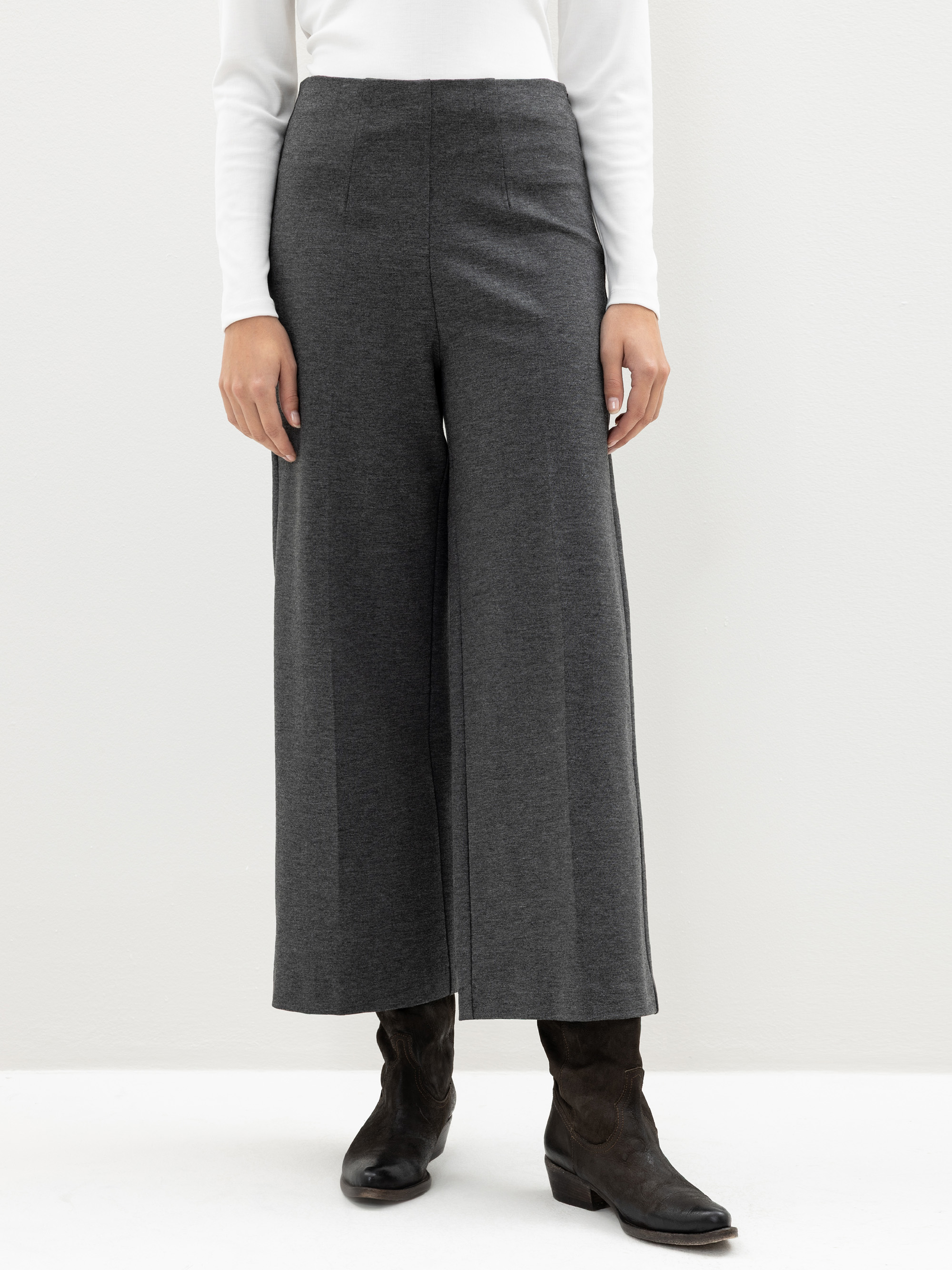 Eine Person, die eine dunkelgraue Hose mit weitem Bein, ein weißes langärmeliges Oberteil und schwarze Stiefel trägt, steht vor einem schlichten hellen Hintergrund. Das Foto ist knapp oberhalb der Schultern abgeschnitten.