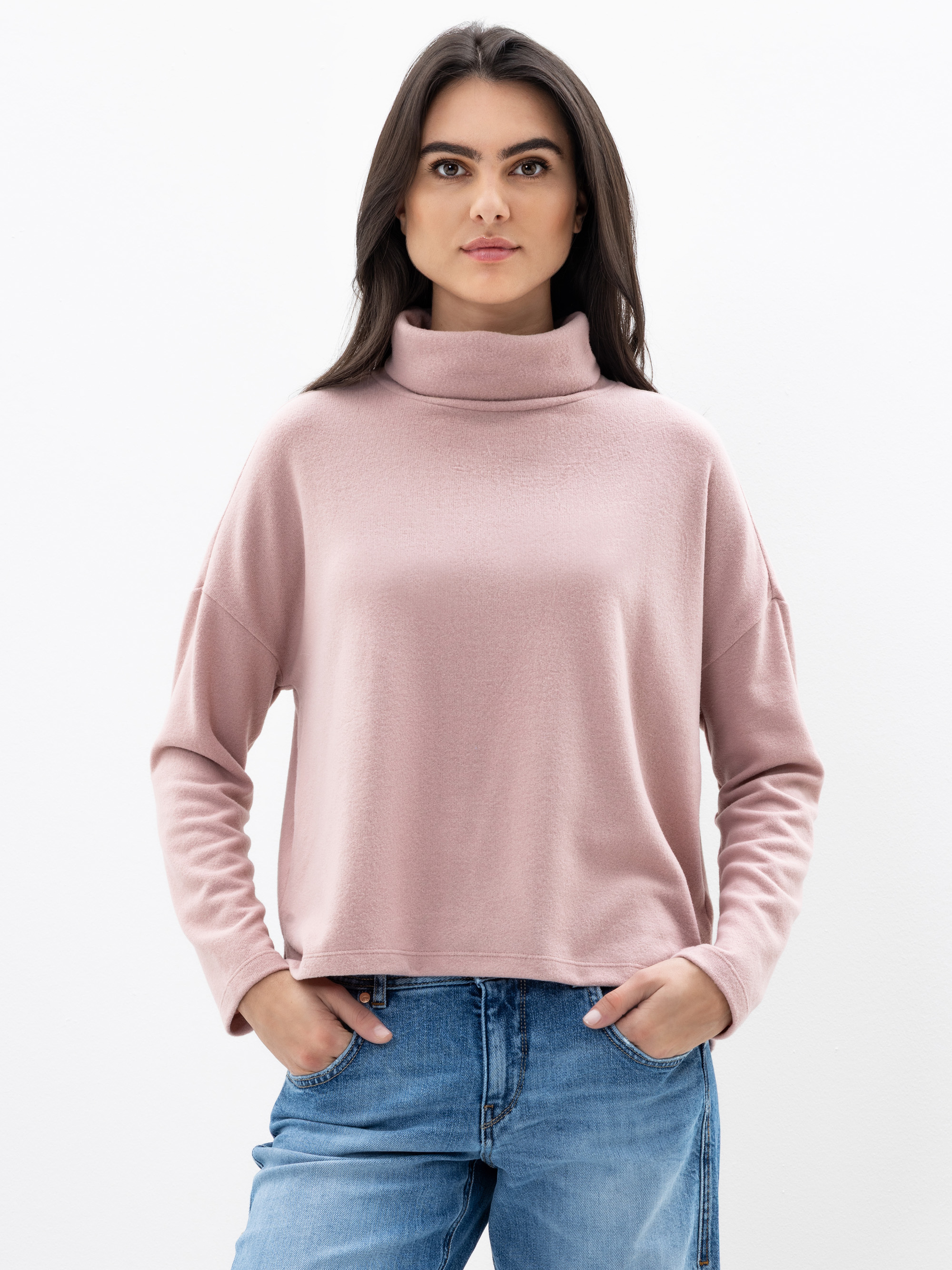 Eine Person steht vor einem schlichten weißen Hintergrund, trägt einen hellrosa Rollkragenpullover und blaue Jeans, hat die Hände in den Taschen und einen neutralen Gesichtsausdruck.