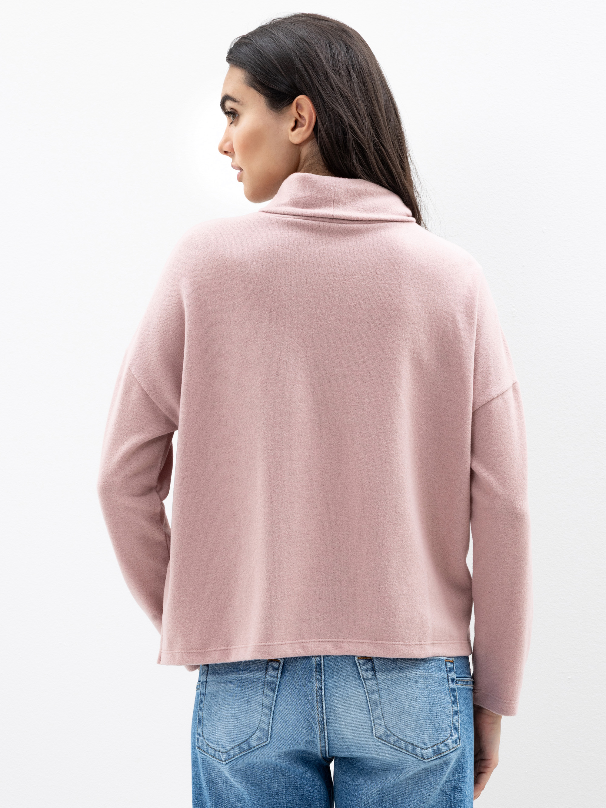 Eine Frau mit langen dunklen Haaren ist von hinten zu sehen. Sie trägt einen hellrosa, langärmeligen, locker sitzenden Pullover mit umgeschlagenem Kragen und dazu eine hellblaue Jeans. Sie steht vor einem schlichten weißen Hintergrund.