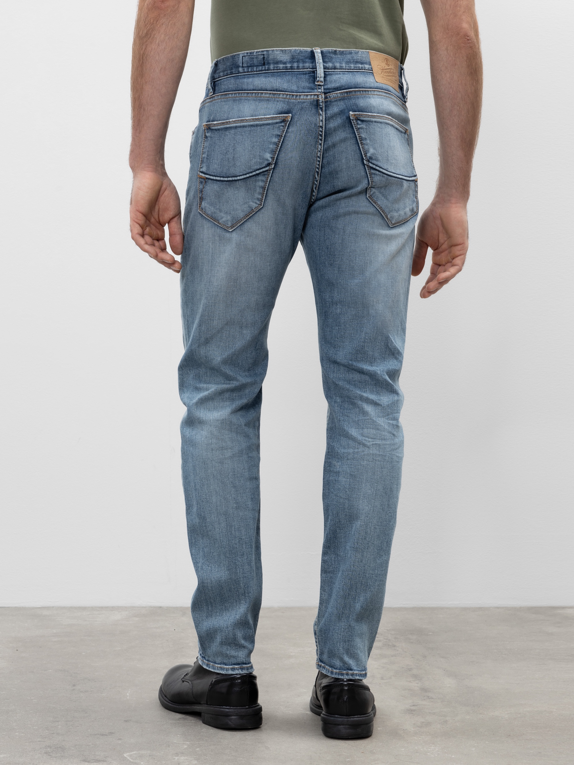 Ein Mann in hellblauen Jeans und schwarzen Schuhen steht mit dem Gesicht zur Kamera und zeigt die Gesäßtaschen der Jeans. Der Hintergrund ist einfarbig und der Boden ist grau.