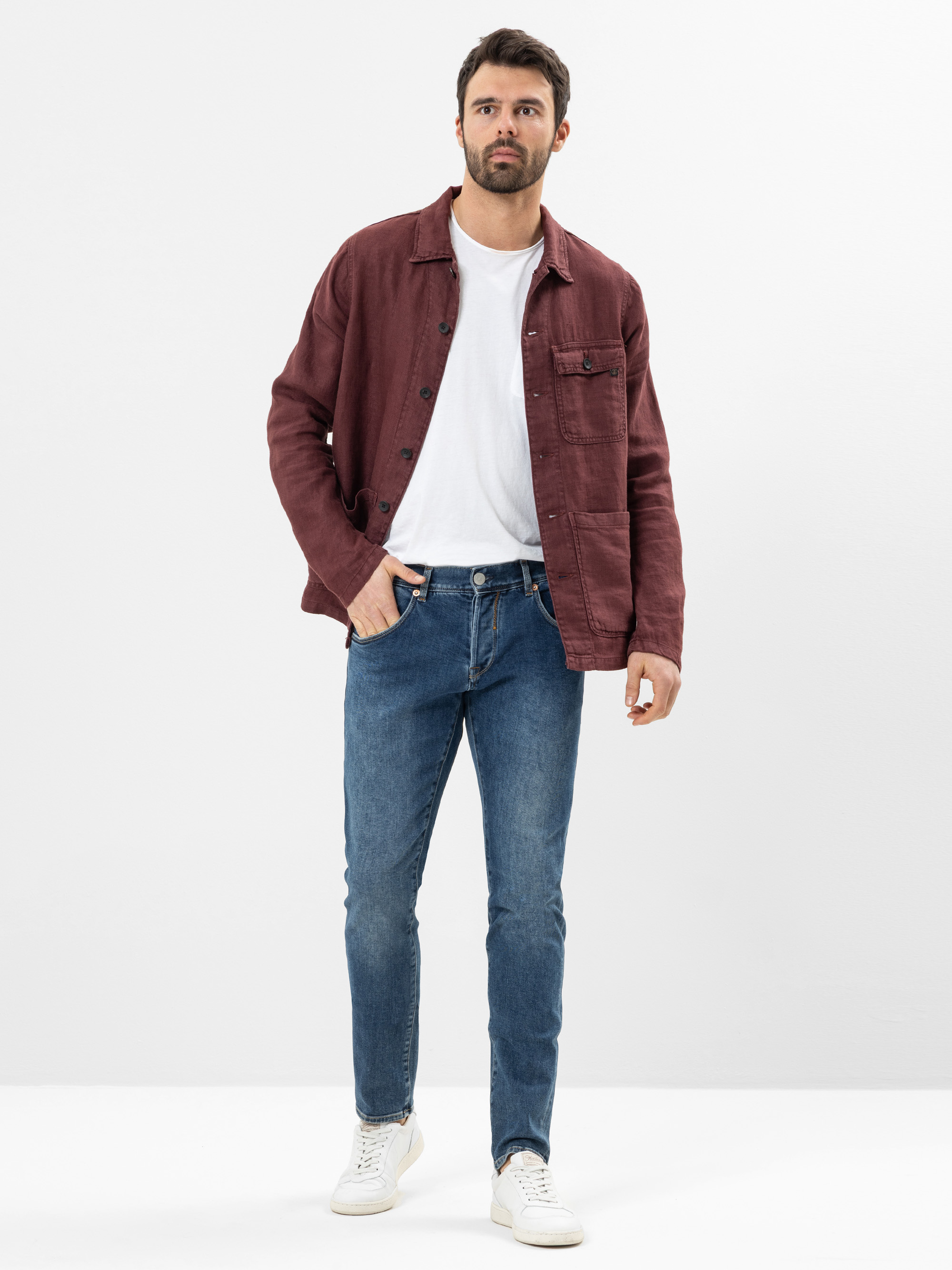 Ein Mann steht vor einem schlichten weißen Hintergrund, trägt eine kastanienbraune Jacke über einem weißen T-Shirt, blaue Jeans und weiße Turnschuhe, hat eine Hand in der Tasche und einen neutralen Gesichtsausdruck.