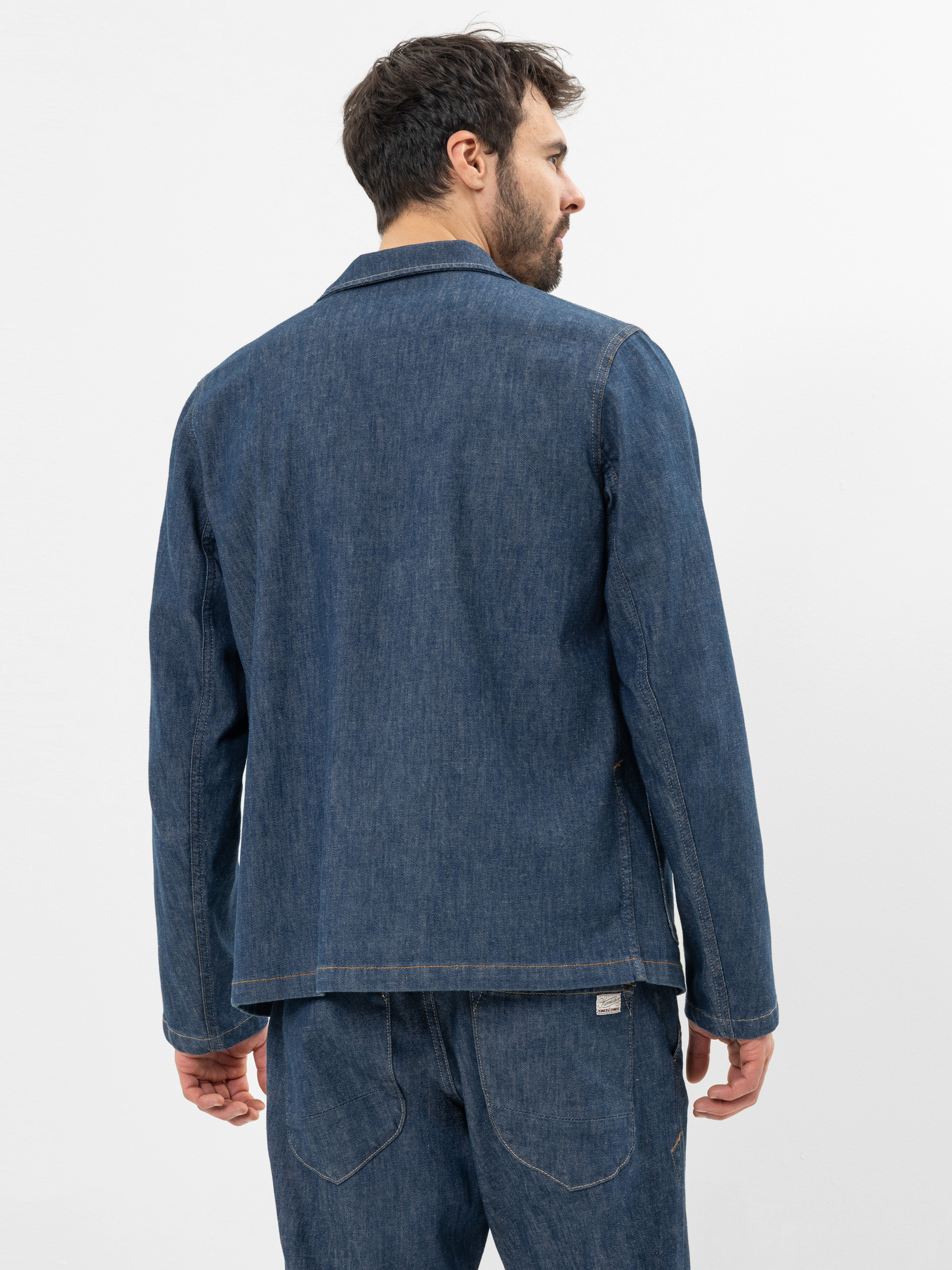 Ein Mann mit dunklem Haar und Bart steht mit dem Rücken zur Kamera und trägt eine blaue Jeansjacke und eine passende Jeanshose vor einem schlichten weißen Hintergrund.