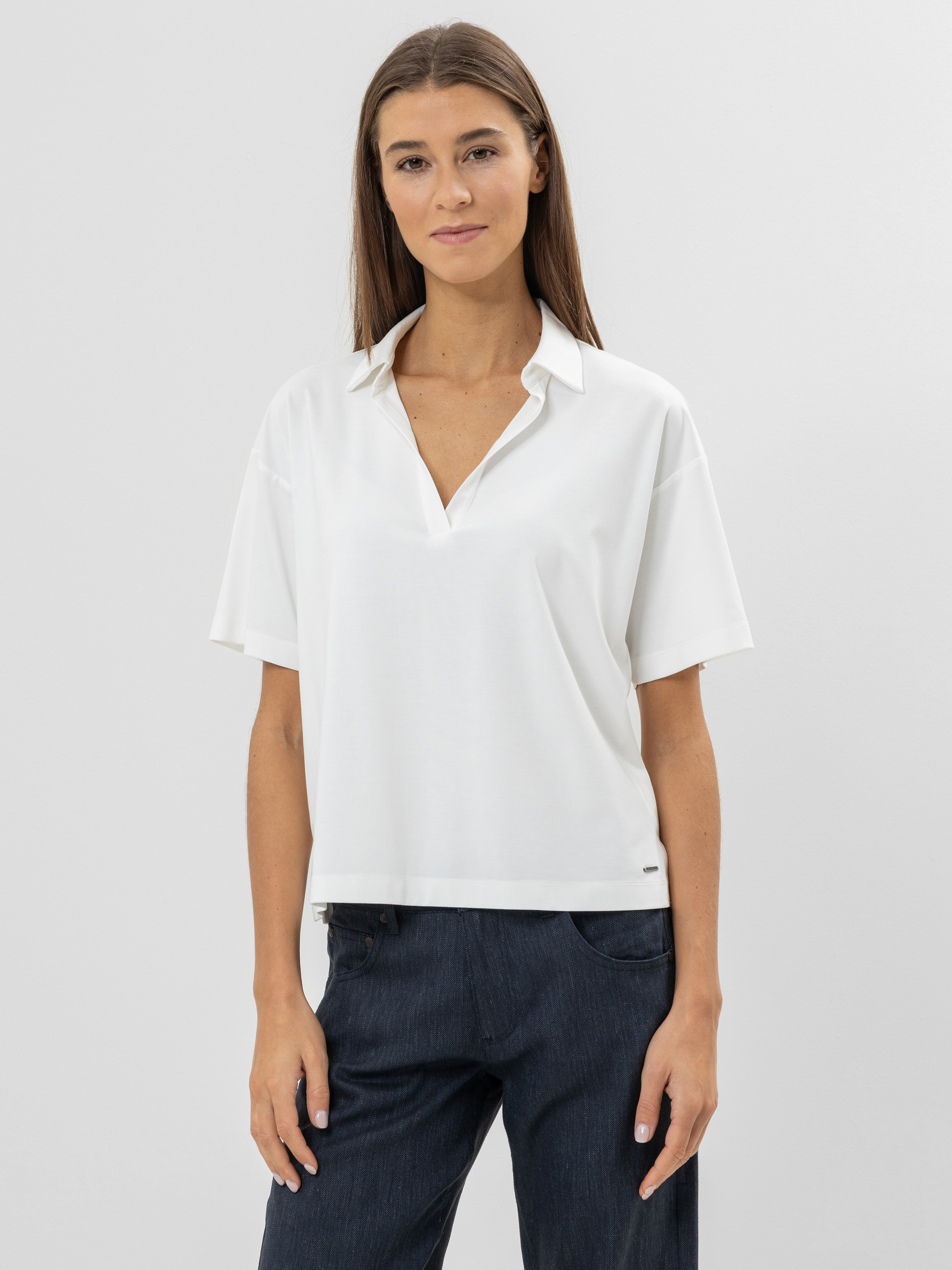 Eine Frau mit glattem braunem Haar, die eine weiße kurzärmelige Bluse mit Kragen und dunkle Jeans trägt, steht mit dem Gesicht zur Kamera vor einem schlichten hellgrauen Hintergrund.
