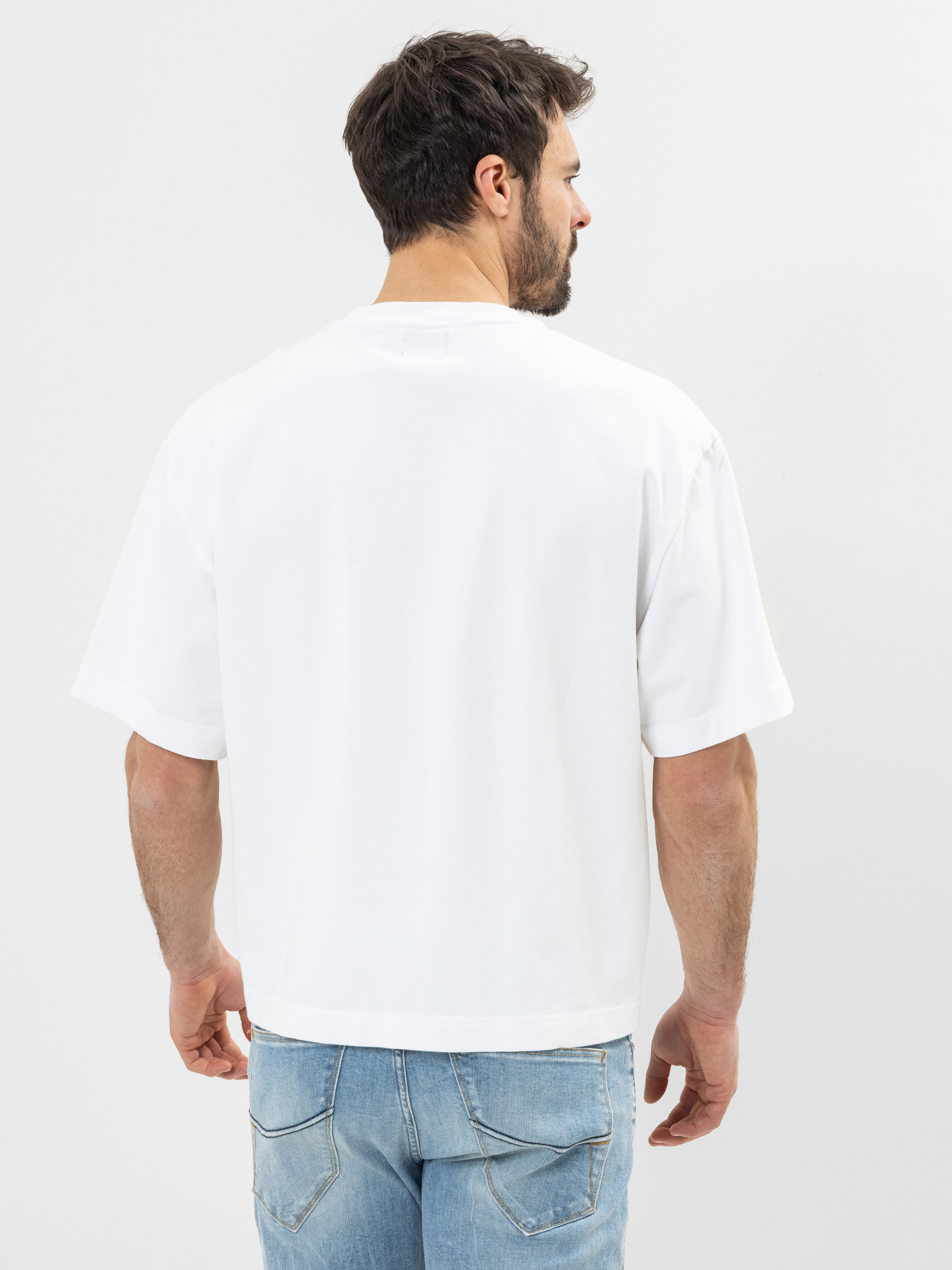 Ein Mann steht mit dem Gesicht zur Wand und trägt ein locker sitzendes weißes kurzärmeliges T-Shirt und hellblaue Jeans. Der Hintergrund ist schlicht und weiß.