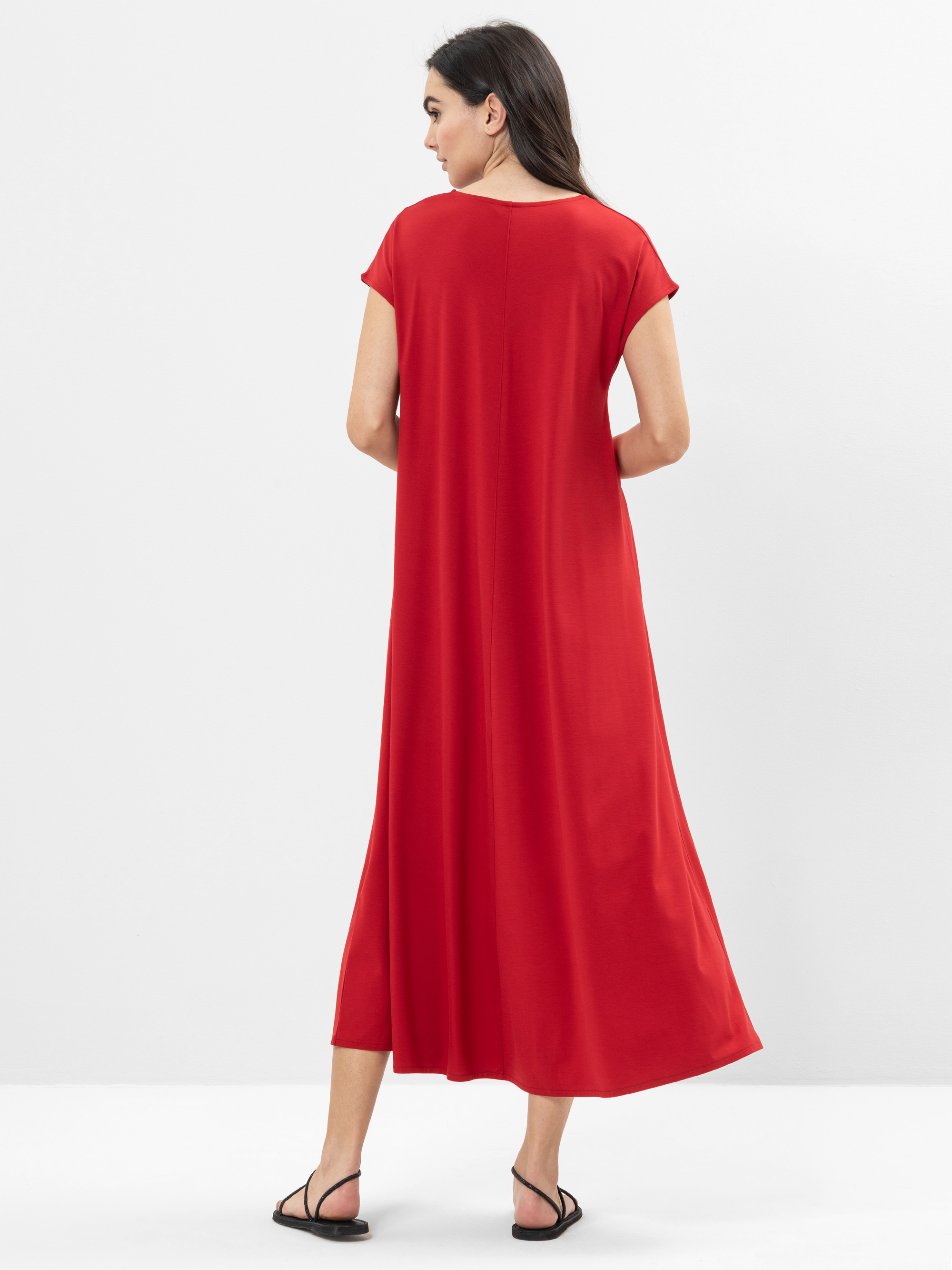 Eine Frau steht mit dem Gesicht zur Wand und trägt ein locker sitzendes, knöchellanges rotes Kleid mit kurzen Ärmeln und schwarzen Sandalen vor einem schlichten weißen Hintergrund.