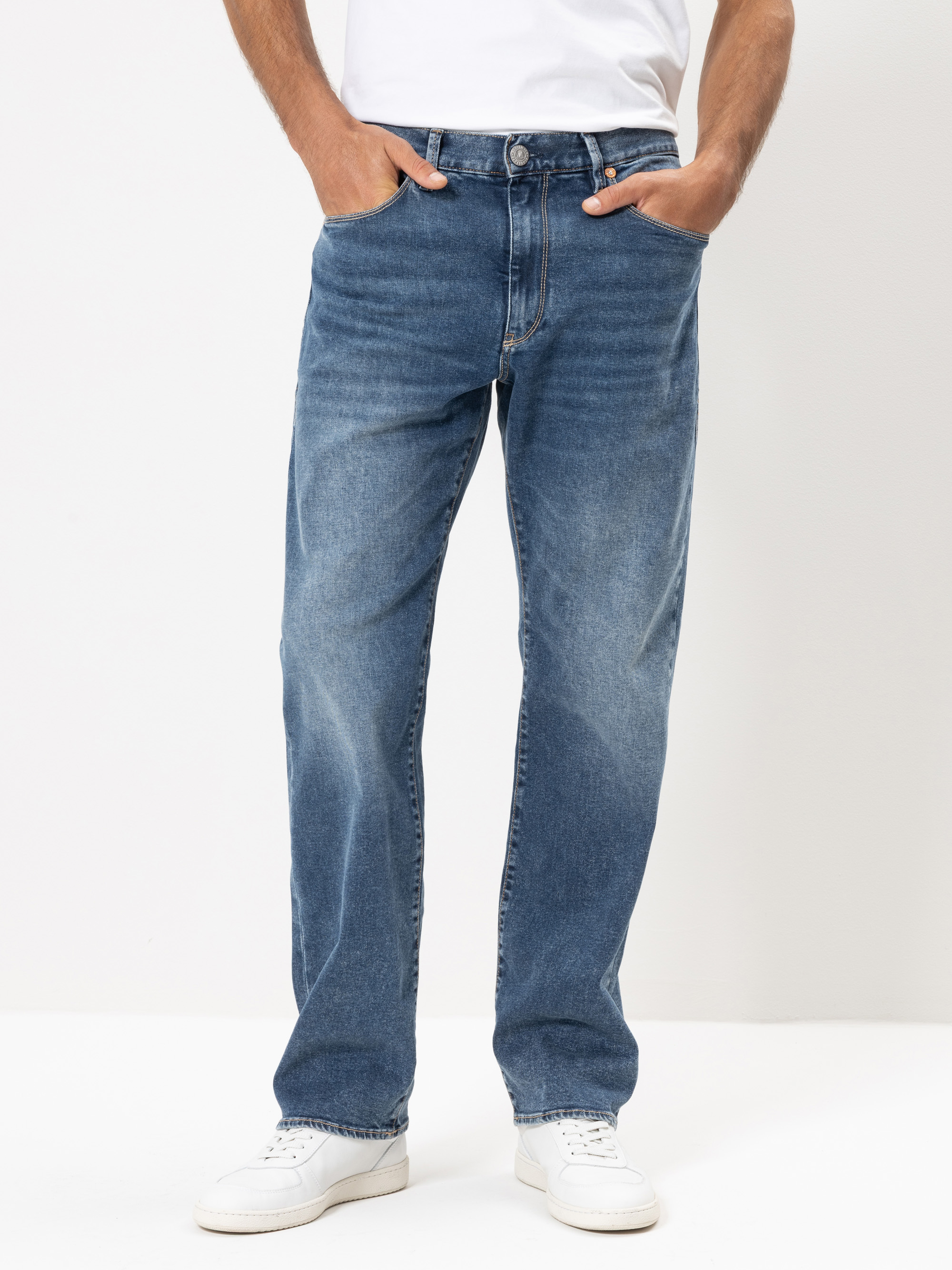 Eine Person in locker sitzenden blauen Jeans und weißen Turnschuhen steht mit den Händen in den Taschen vor einem schlichten weißen Hintergrund. Der Oberkörper ist teilweise sichtbar.