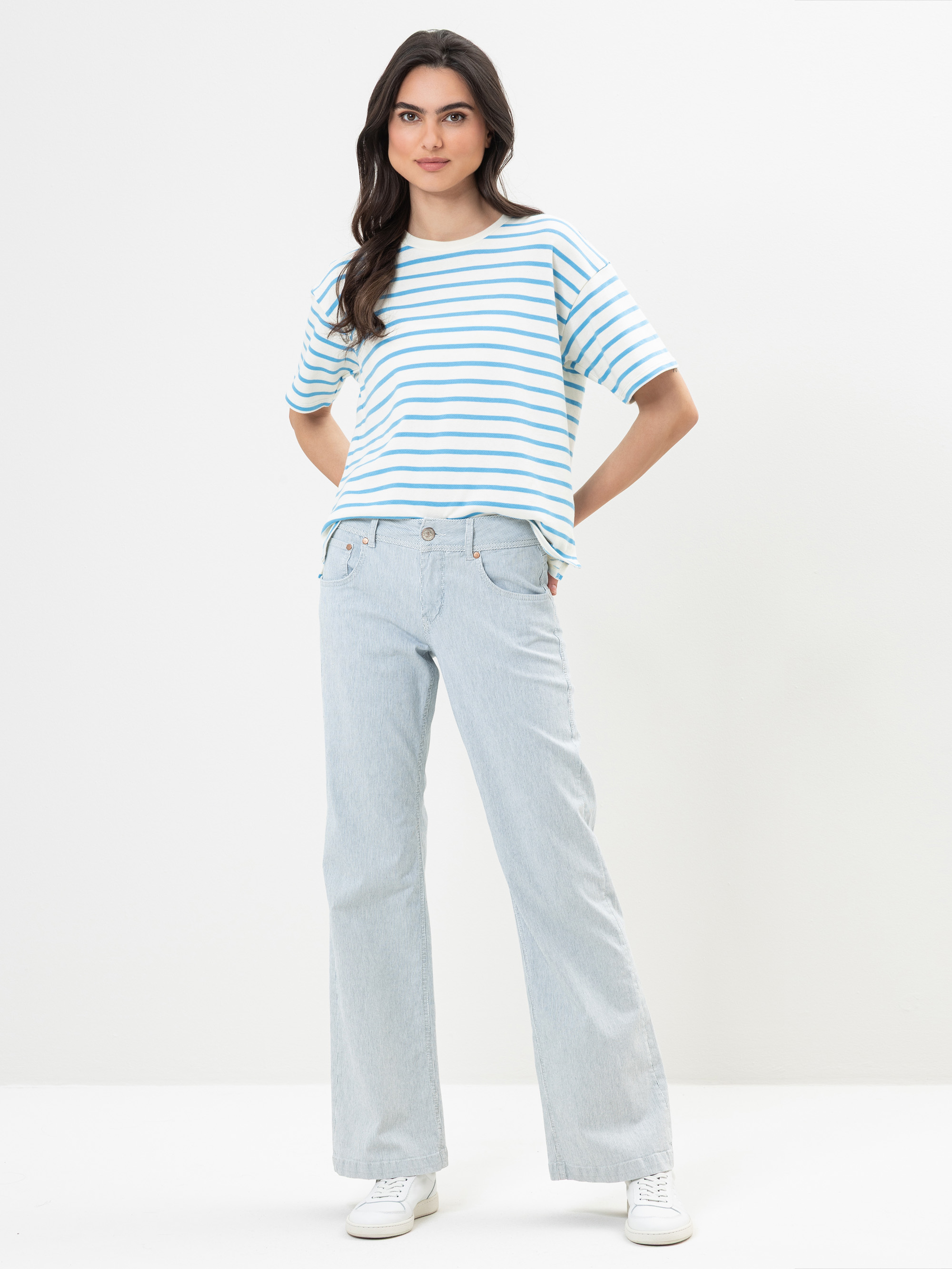 Eine Frau steht vor einem schlichten weißen Hintergrund und trägt ein blau-weiß gestreiftes T-Shirt, hellblaue Jeans und weiße Turnschuhe. Sie hat langes, dunkles Haar und verschränkt leicht lächelnd die Hände hinter dem Rücken.