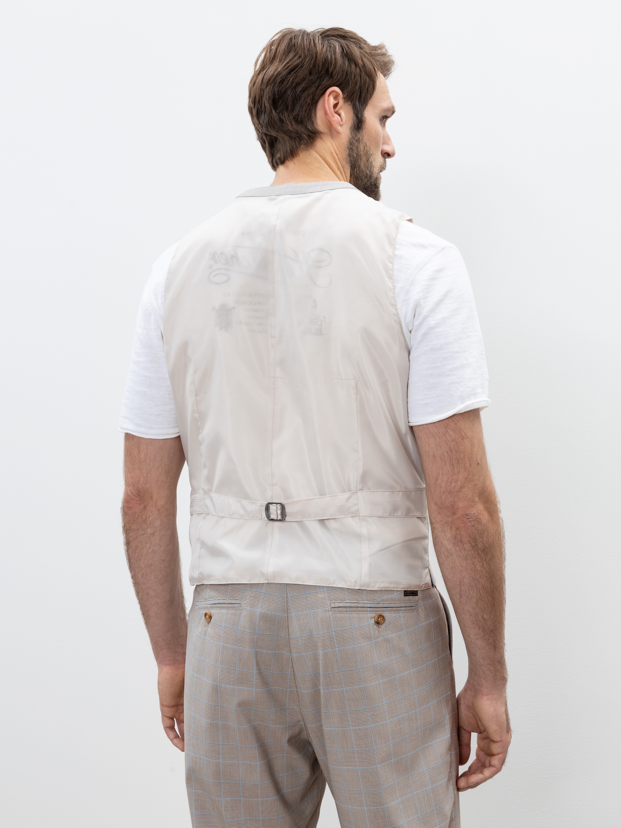 Ein Mann mit hellbraunem Haar ist von hinten zu sehen. Er trägt ein weißes kurzärmeliges Hemd, eine hellbeige Weste mit verstellbarem Gürtel und eine grau karierte Hose vor einem einfarbig weißen Hintergrund.