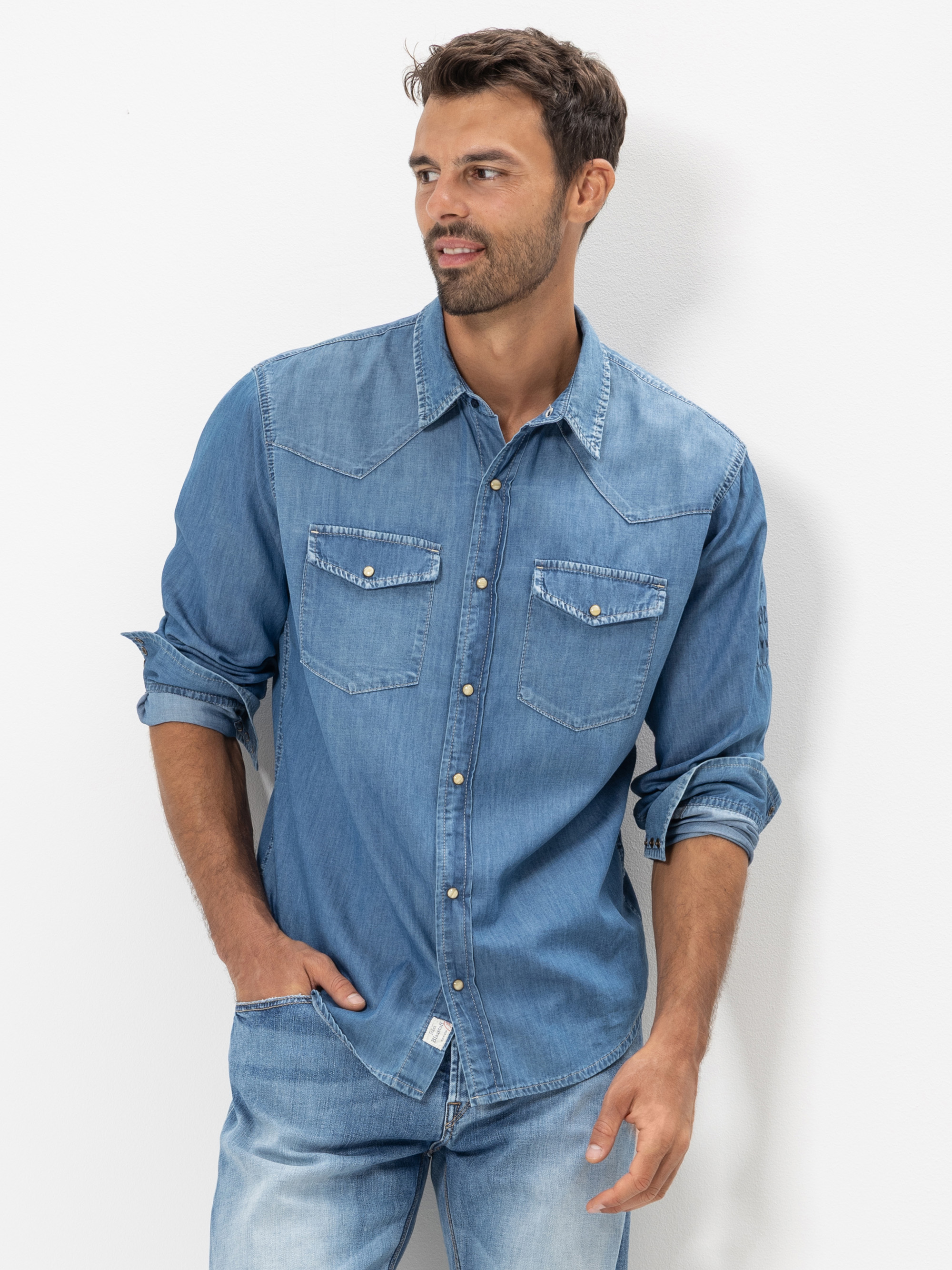 Ein Mann mit kurzen dunklen Haaren und einem Bart trägt ein langärmeliges Jeanshemd mit Perlmuttknöpfen und Vordertaschen. Er steht mit einer Hand in der Tasche seiner Jeans und blickt zur Seite vor einem schlichten weißen Hintergrund.