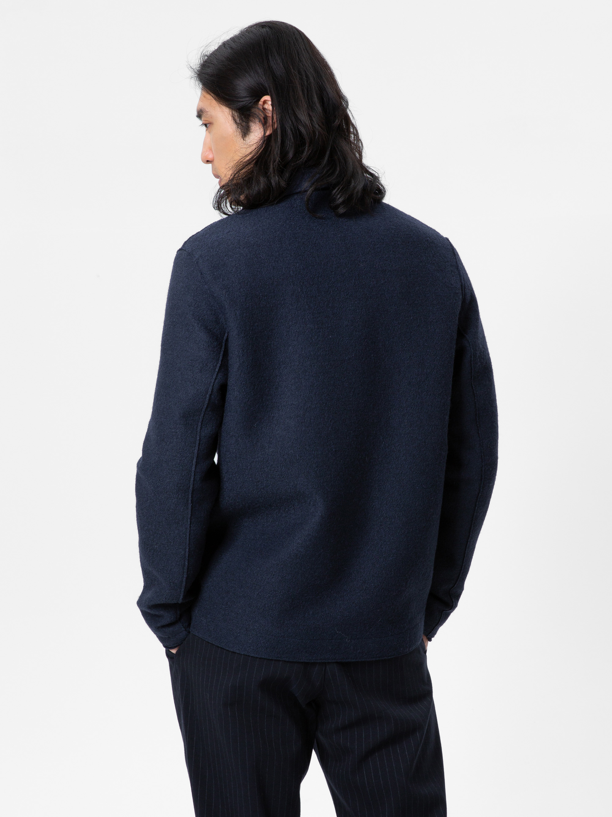 Ein Mann mit langen dunklen Haaren, der eine marineblaue langärmelige Jacke und eine dunkle Nadelstreifenhose trägt, steht mit dem Gesicht zur Kamera vor einem schlichten weißen Hintergrund.