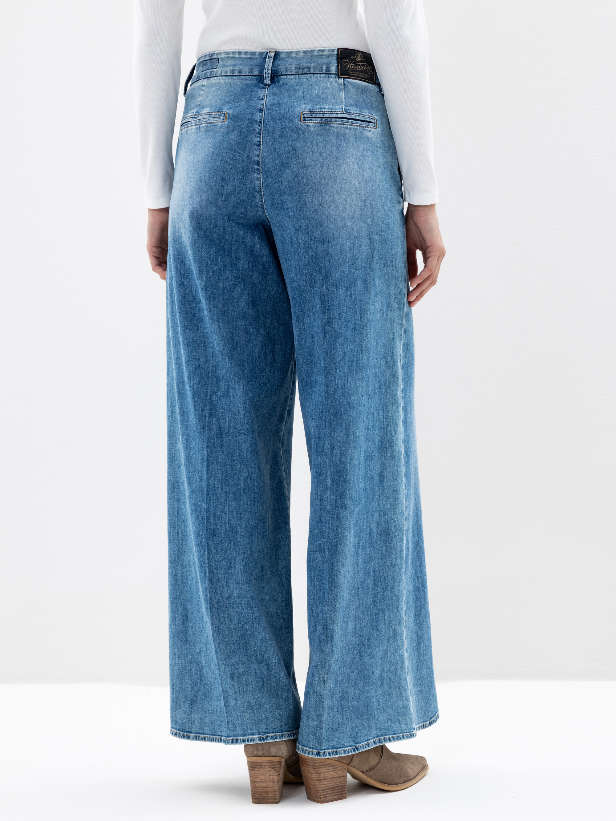 Eine Person trägt eine blaue Jeans mit weitem Bein und hellbraune Schuhe mit Absätzen, von hinten gesehen. Die Jeans hat Gesäßtaschen und ein dunkles Etikett am Bund. Die Person trägt außerdem ein weißes langärmeliges Oberteil.
