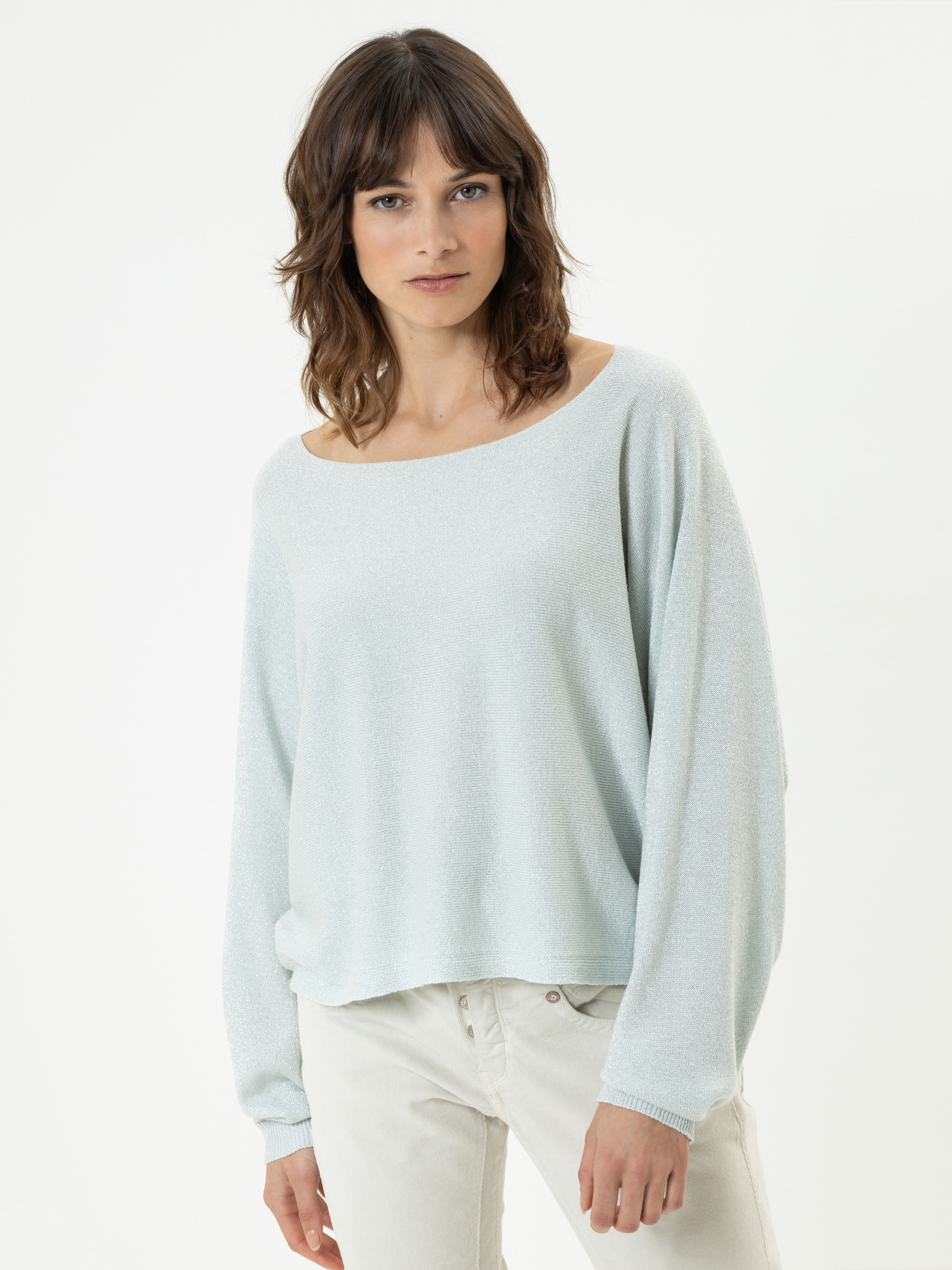 Eine Frau mit schulterlangen braunen Haaren trägt einen hellblauen weiten Pullover und eine helle Hose, steht vor einem schlichten weißen Hintergrund und blickt in die Kamera.