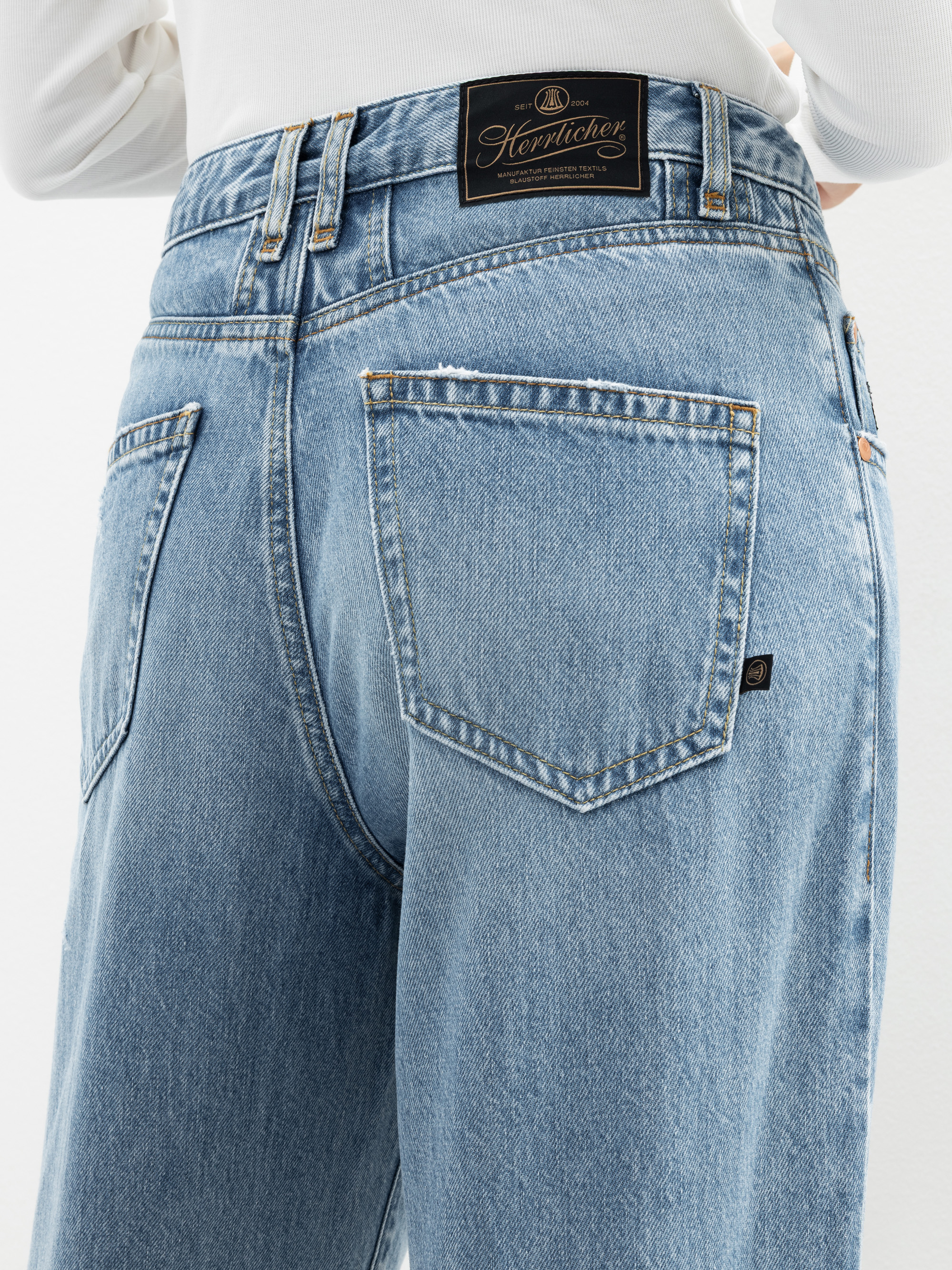 Nahaufnahme einer Person, die eine hellblaue Jeans mit hoher Taille, einem schwarzen Herrlicher-Markenaufnäher am Bund und zwei Gesäßtaschen trägt, dazu ein weißes langärmeliges Oberteil.