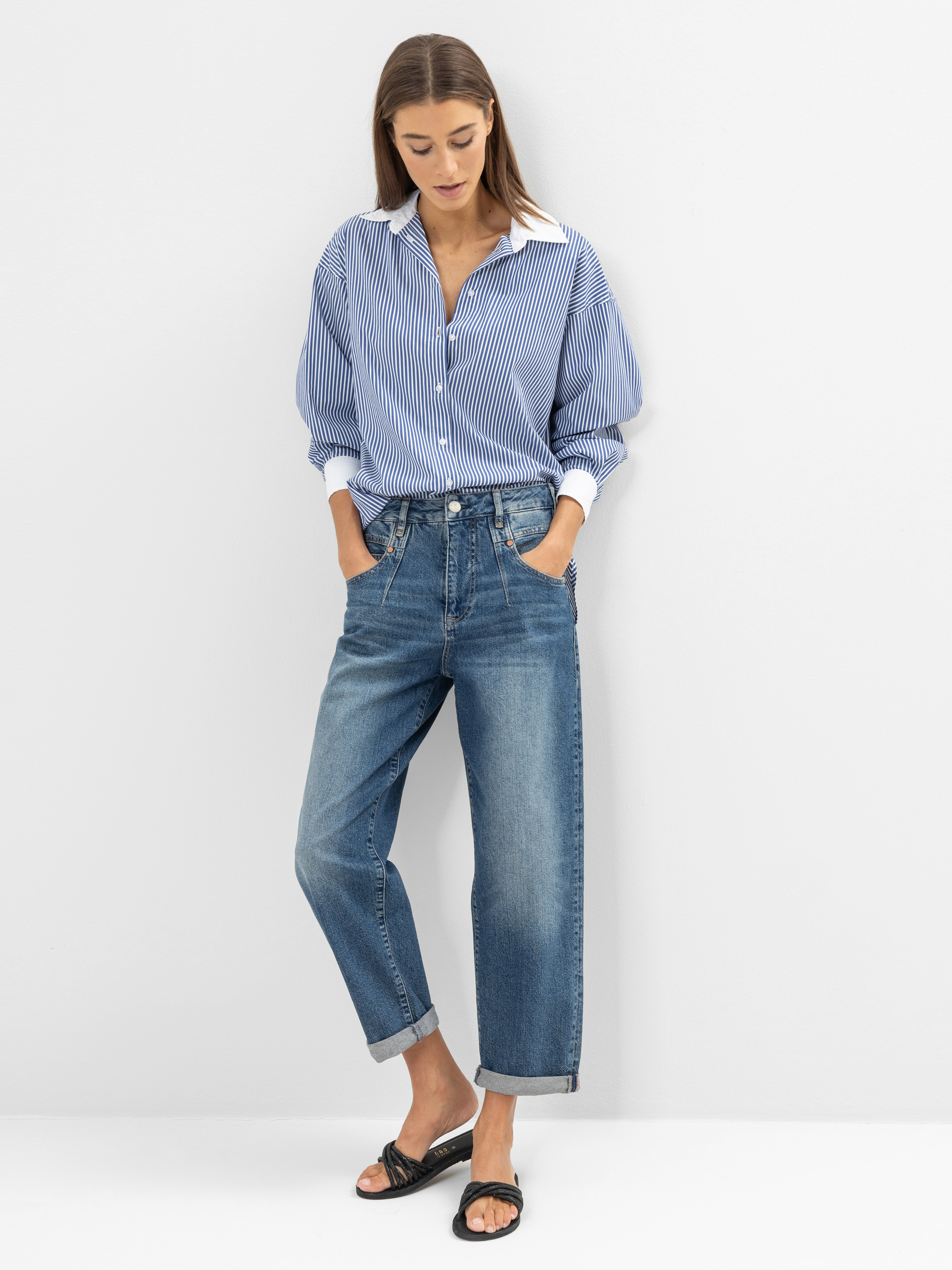 Eine Frau steht vor einer weißen Wand. Sie trägt ein blau-weiß gestreiftes Hemd mit hochgekrempelten Ärmeln, eine blaue Jeans mit hoher Taille und Manschetten und schwarze, offene Sandalen. Ihre Hände stecken in den Taschen und ihr Kopf ist nach unte