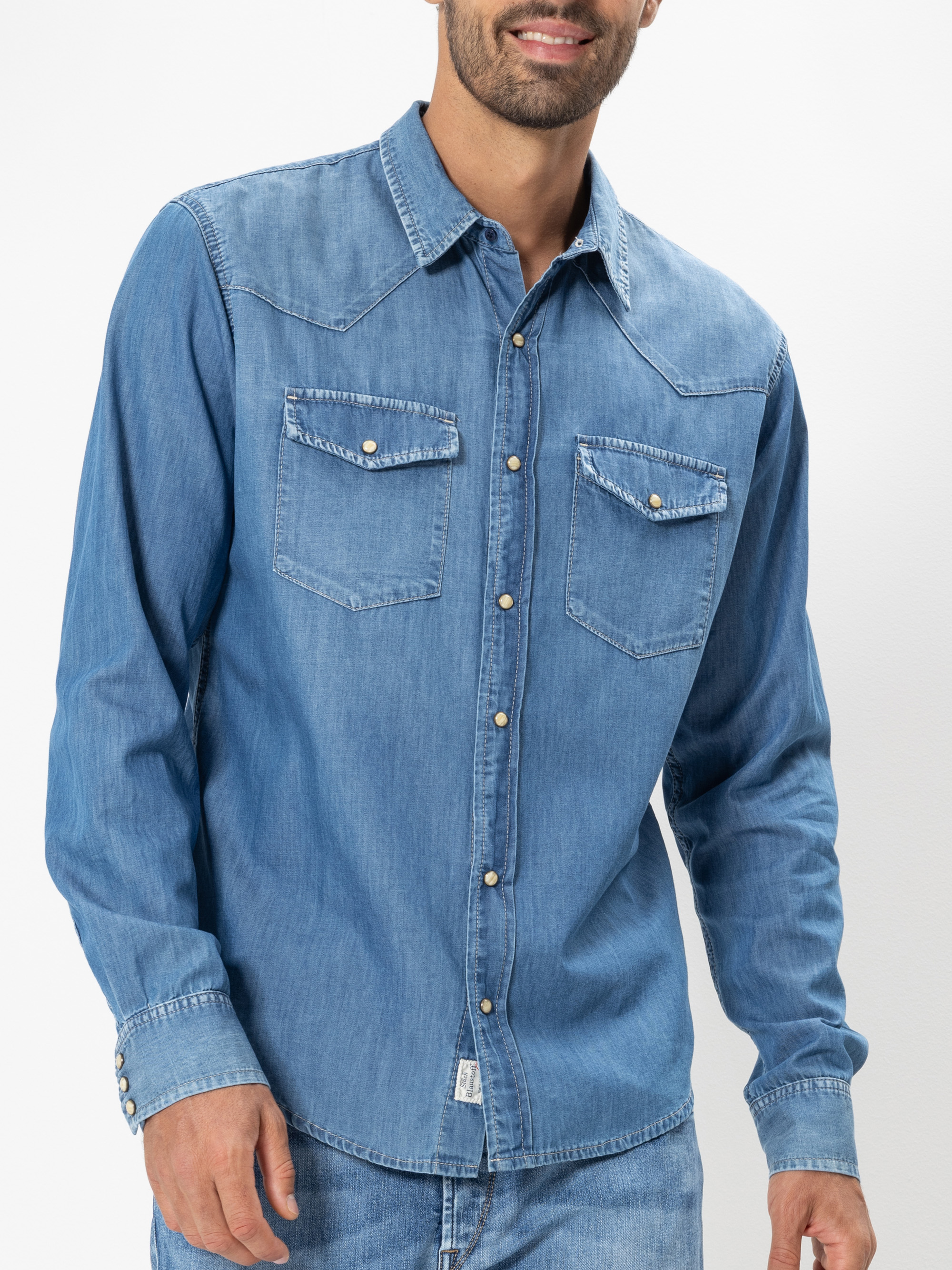 Ein Mann, der ein hellblaues Jeanshemd mit Druckknöpfen und Klappentaschen auf der Brust trägt, gepaart mit einer blauen Jeans, steht vor einem schlichten weißen Hintergrund.