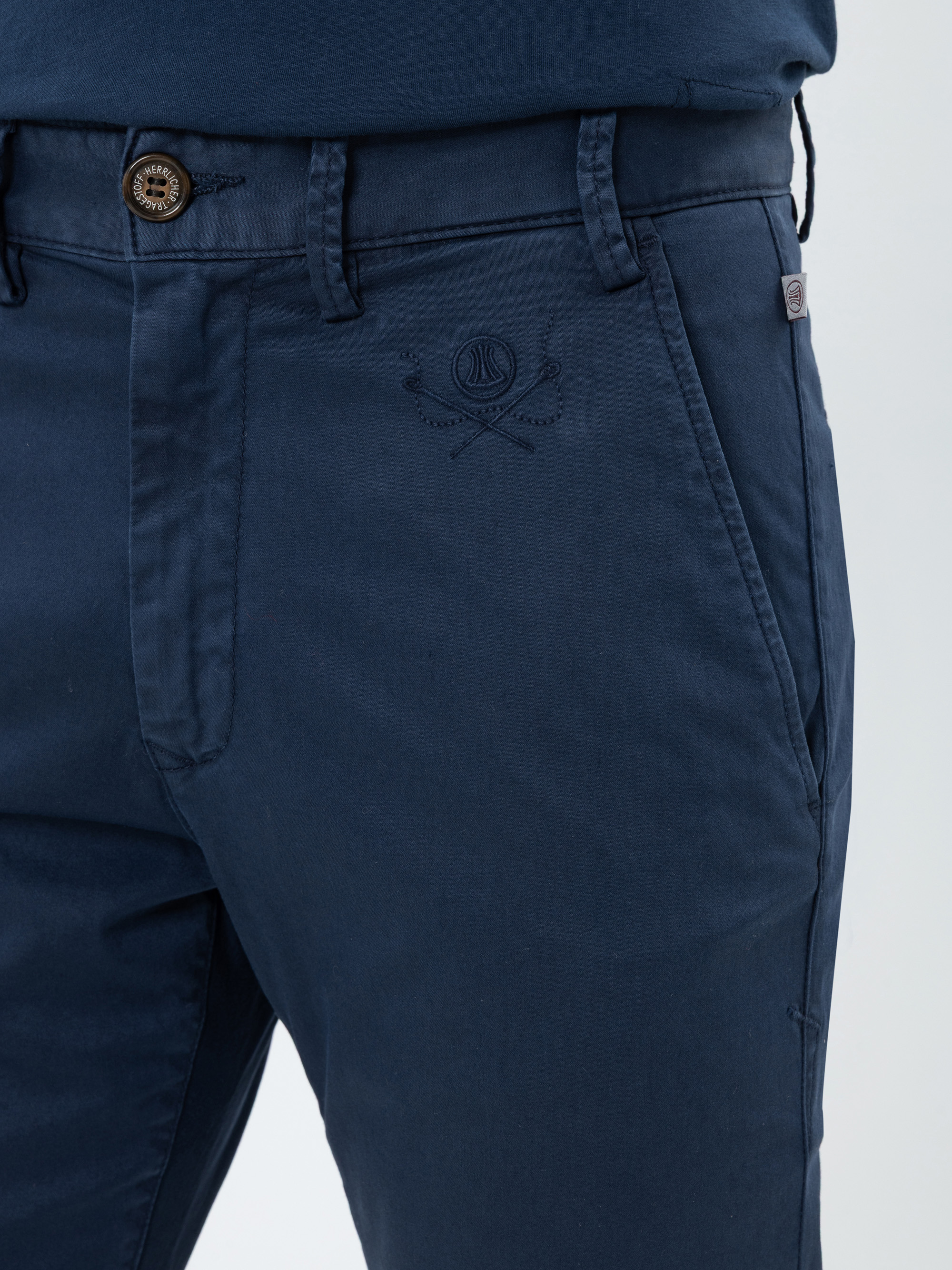 Nahaufnahme einer Person, die eine dunkelblaue Hose mit Knopf und Gürtelschlaufen trägt, auf der über der rechten Tasche ein dezentes gesticktes Tennislogo zu sehen ist. Das marineblaue Hemd der Person ist teilweise sichtbar.