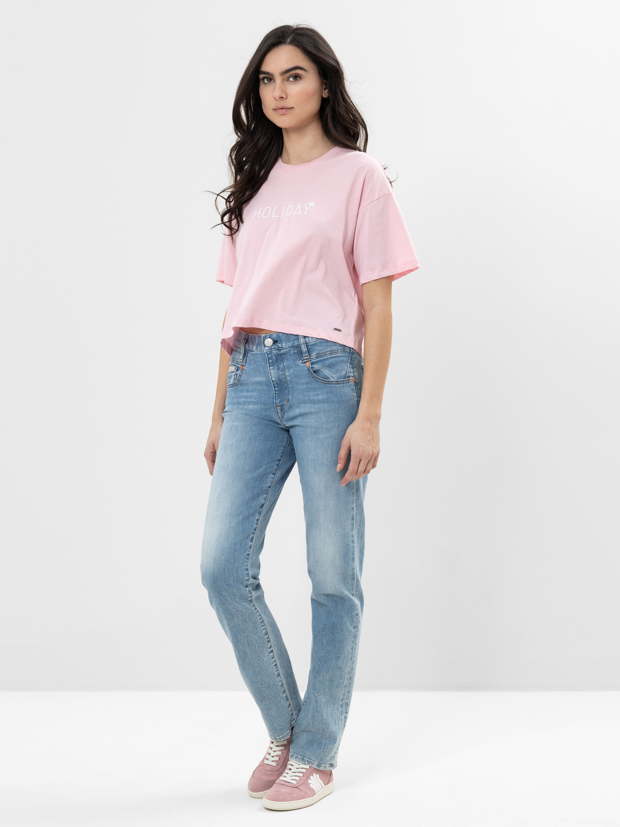 Eine Frau mit langen dunklen Haaren, die ein lockeres rosa T-Shirt, hellblaue Jeans und rosa Turnschuhe trägt, steht vor einem schlichten hellgrauen Hintergrund und schaut mit neutralem Blick in die Kamera.