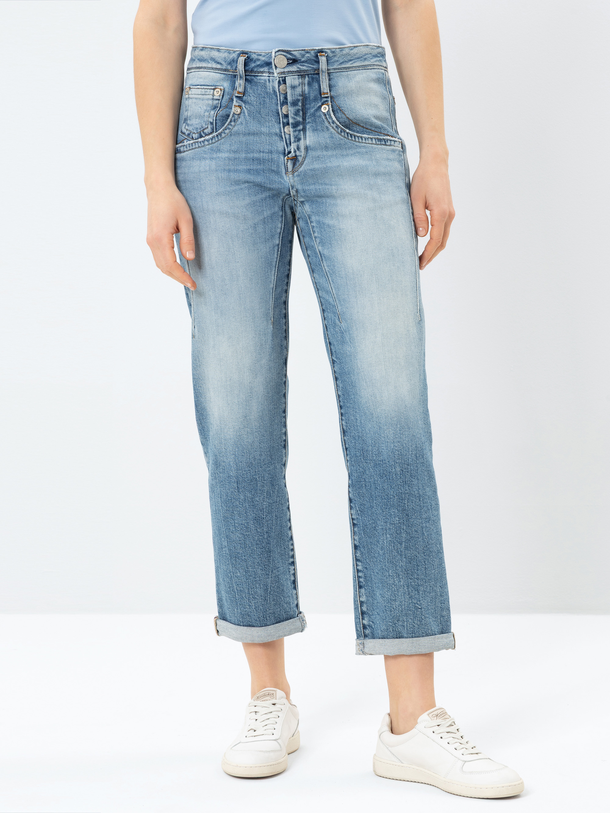 Person, die eine hellblaue, leicht verblichene Jeans mit geradem Bein und an den Knöcheln aufgerollten Manschetten trägt, gepaart mit weißen Turnschuhen und einem hellblauen Oberteil, stehend vor einem schlichten weißen Hintergrund.
