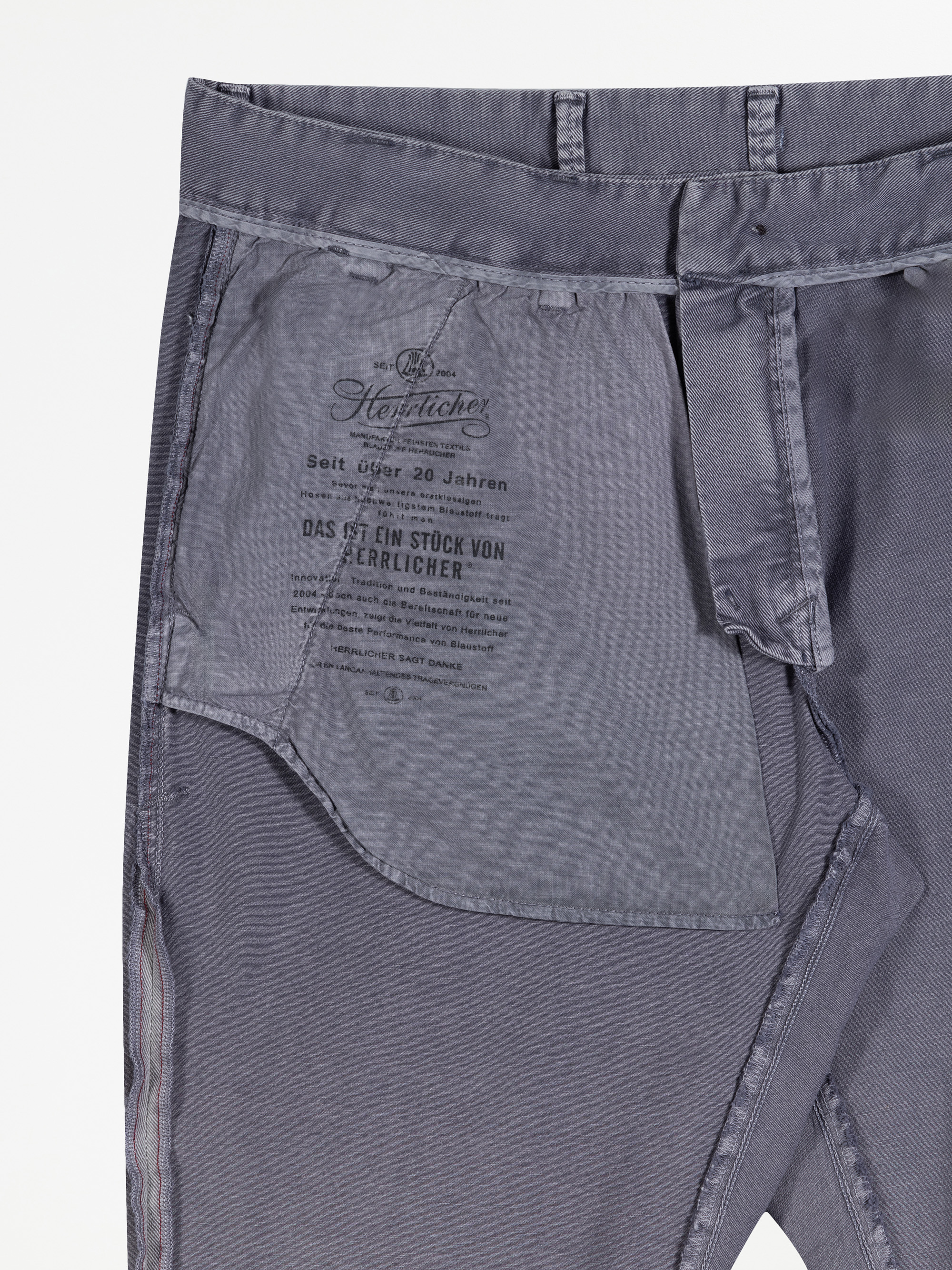 Nahaufnahme der Innenseite einer grauen Hose, die das Innenfutter der Taschen mit aufgedrucktem schwarzem Text in deutscher Sprache und einem stilisierten Logo in der Nähe des Hosenbundes zeigt.