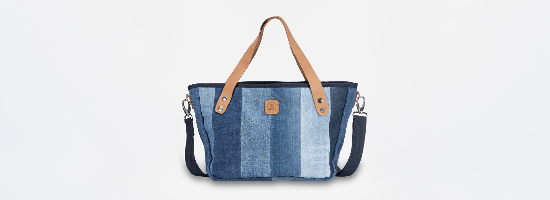 Eine Tragetasche aus vertikalem Patchwork-Denim in verschiedenen Blautönen, mit hellbraunen Ledergriffen, einem abnehmbaren dunklen Riemen und einem kleinen braunen Logoaufnäher auf der Vorderseite vor weißem Hintergrund.