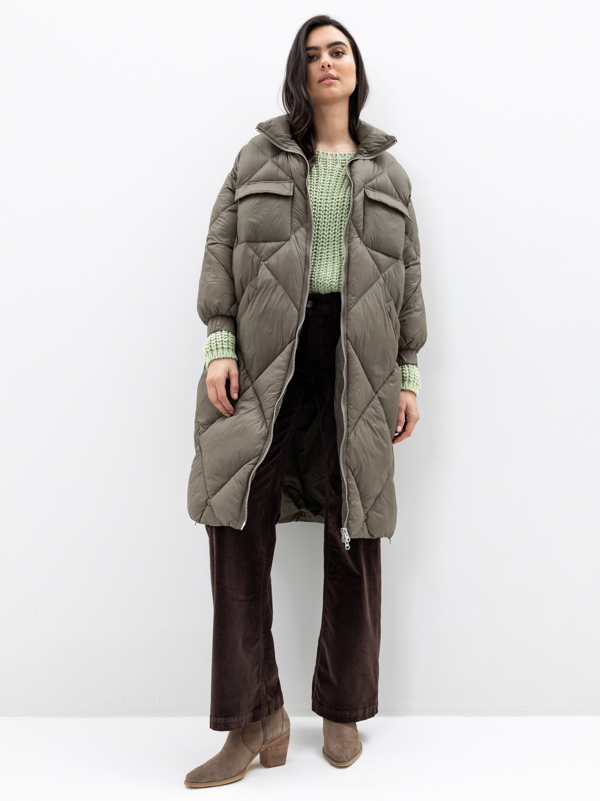 Eine Frau steht vor einem weißen Hintergrund und trägt einen langen olivgrünen Puffermantel, einen hellgrünen Strickpullover, eine braune Cordhose und hellbraune Stiefel mit Absatz. Sie hat langes dunkles Haar und einen neutralen Gesichtsausdruck.