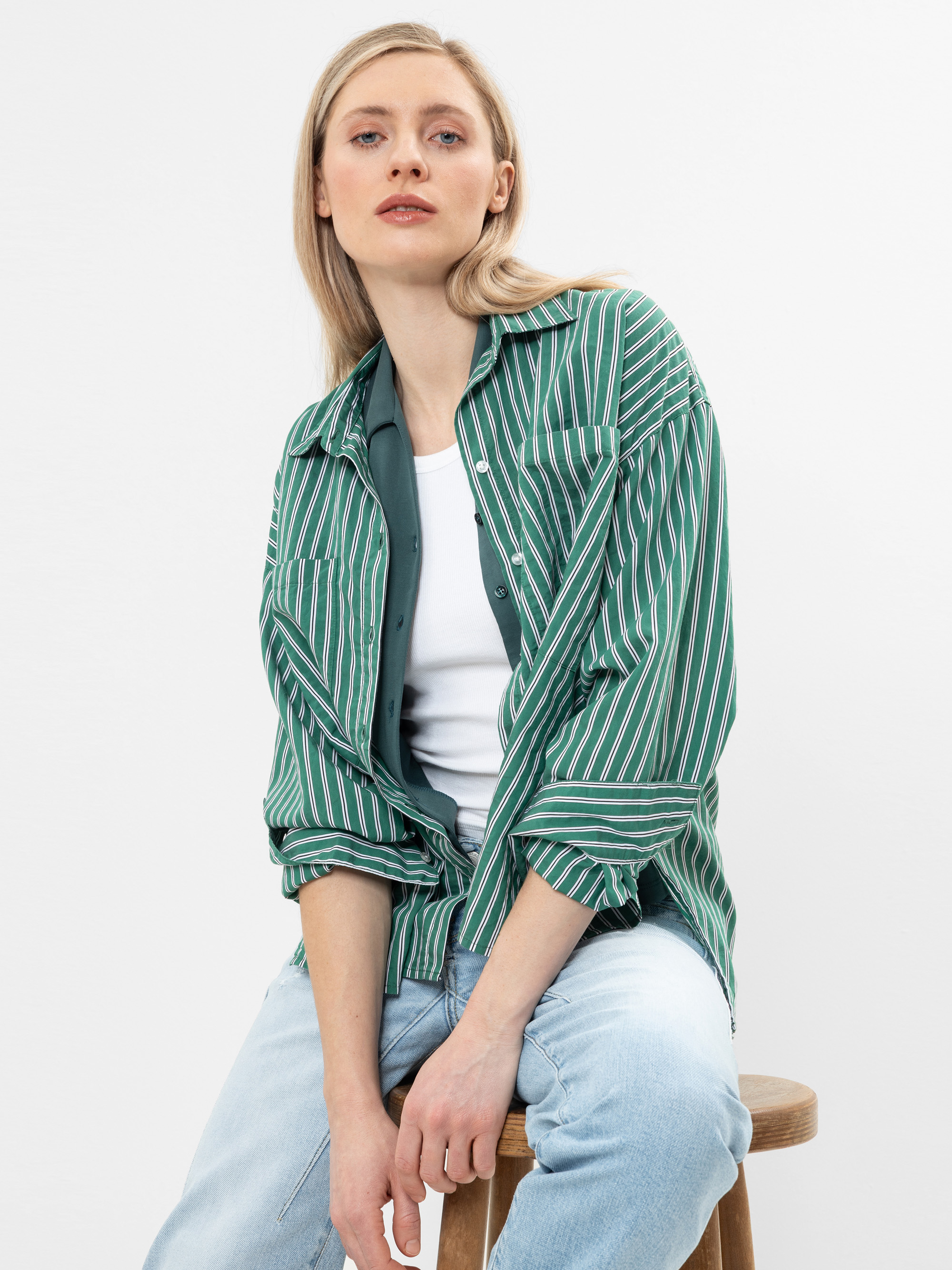 Eine Person mit langen blonden Haaren, die ein grün-weiß gestreiftes Hemd über einem weißen Top und hellblaue Jeans trägt, sitzt auf einem Holzhocker vor einem schlichten weißen Hintergrund.