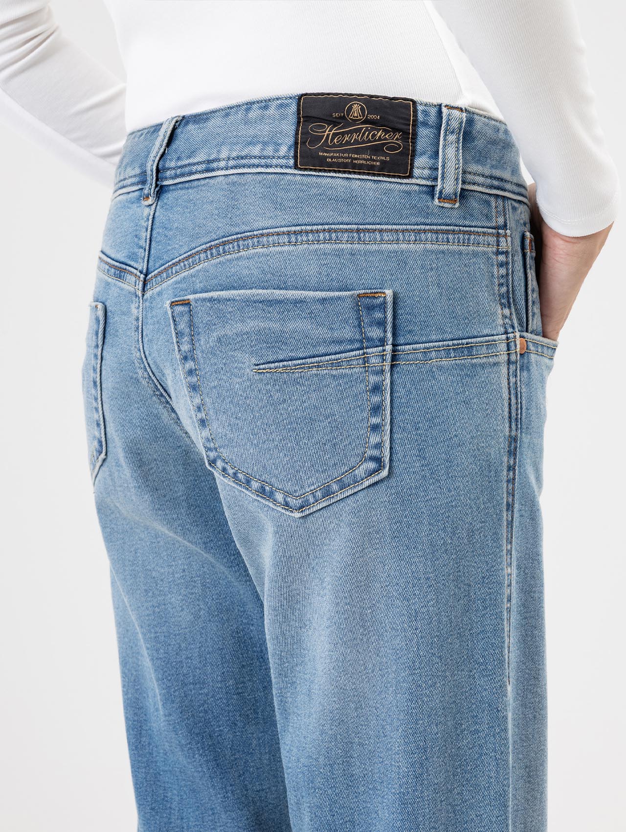 Eine Person trägt eine hellblaue, hochtaillierte Jeans und ein weißes, langärmeliges Oberteil, von hinten gesehen. Die Jeans hat einen schwarzen Lederaufnäher mit goldenem Hemitique-Branding am Bund.